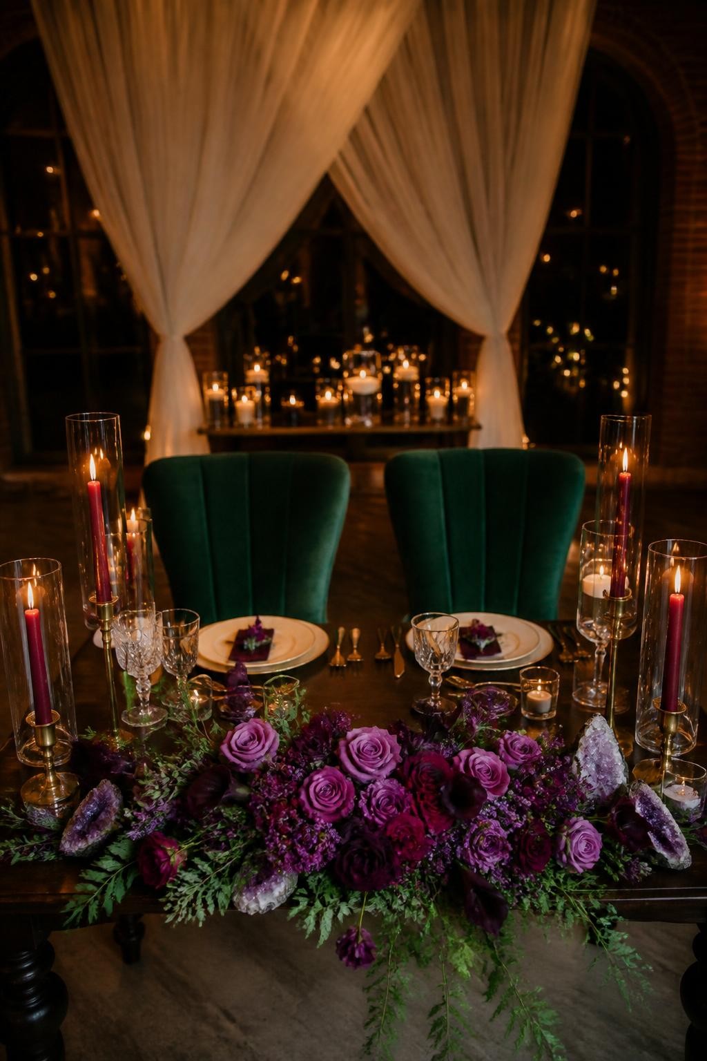 Realistic DSLR photo of a fall wedding sweetheart table with emerald velvet chairs, amethyst floral arrangement, ruby taper candles, gold flatware, cream plates, dark wood table, soft draped backdrop, warm candlelight, romantic jewel tone reception setup.