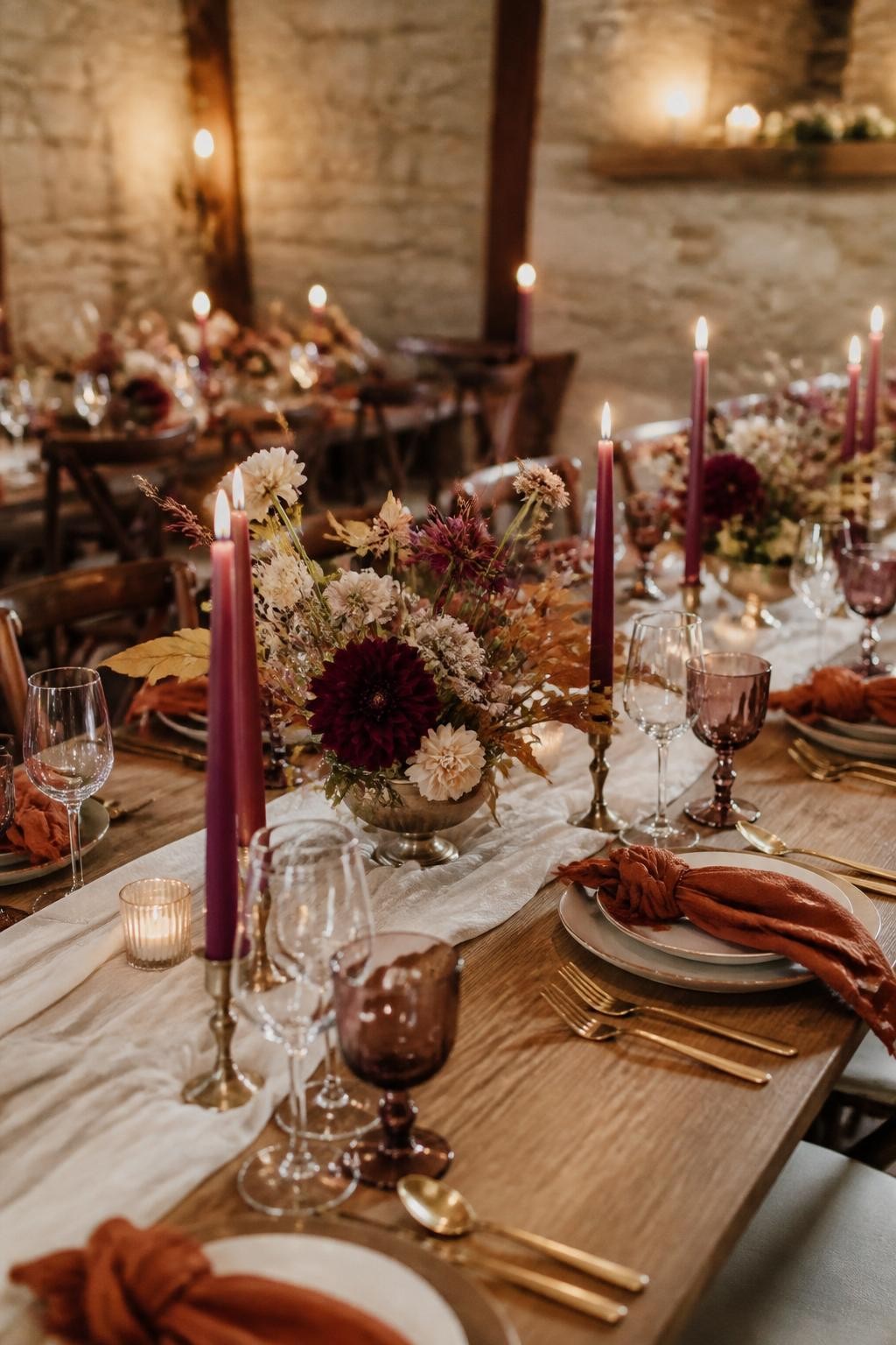 Realistic DSLR wedding reception photo inside a stone barn with long wooden tables, burnt sienna napkins, fig purple taper candles, antique gold flatware, ivory linen runners, low floral centerpieces with dahlias, scabiosa, dried grasses, and soft candlelight, shallow depth of field, warm fall evening mood, no visible faces.