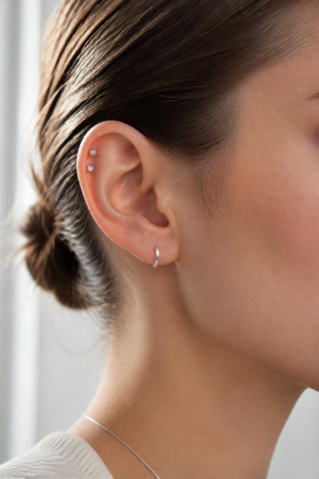 Side portrait of an ear with neutral fair skin tone, two tiny opal studs placed neatly on the forward helix near the top of the ear, sleek pulled-back hair, minimal silver jewelry, soft window light, realistic beauty editorial style, clear focus on the forward helix area, clean skin texture.