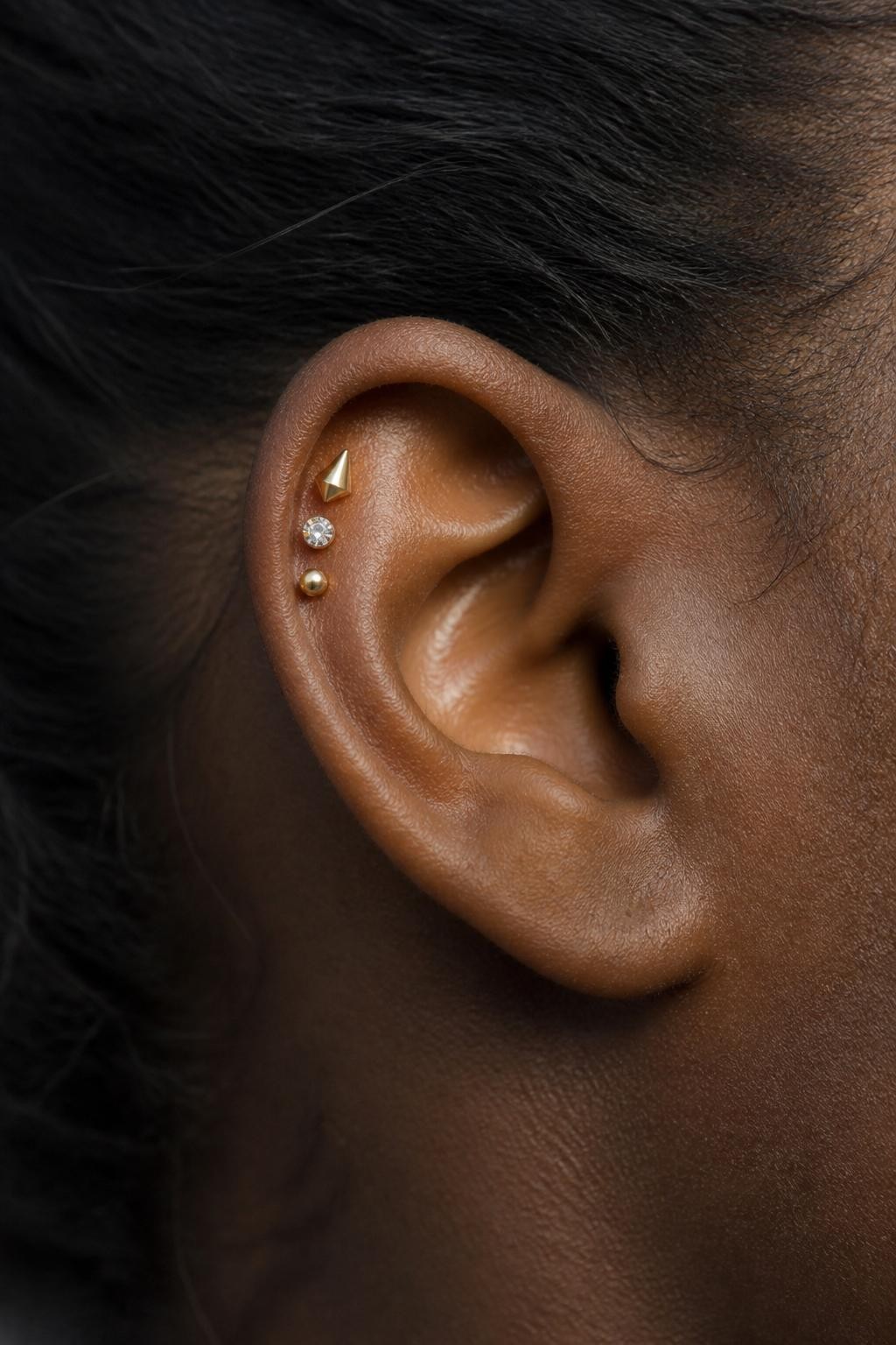 Close-up side portrait of an ear with deep brown skin tone, flat upper cartilage decorated with a tiny gold spike stud, one small crystal stud, and one tiny round gold dot, arranged in a compact cluster, hair pulled away from ear, natural daylight, realistic DSLR editorial photo.
