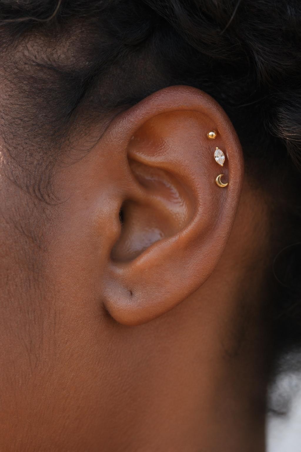 Close-up side portrait of an ear with rich dark skin tone, upper helix styled with three different tiny earrings: one gold ball stud, one small marquise crystal, one tiny moon-shaped stud, uneven but balanced placement, natural skin texture, soft daylight, realistic DSLR beauty photography.
