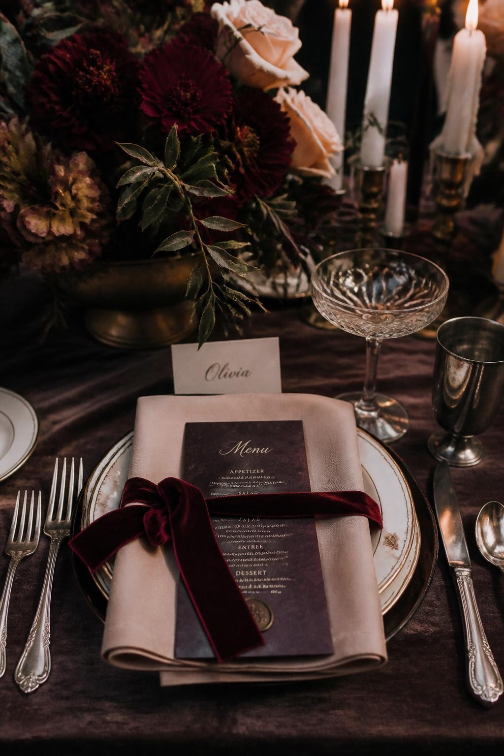 Realistic DSLR photo of a fall wedding reception place setting with garnet velvet ribbon, blush beige linen napkin, smoked plum menu card, old silver flatware, crystal coupe glass, dark floral centerpiece, candle wax drips, moody luxury wedding detail shot.