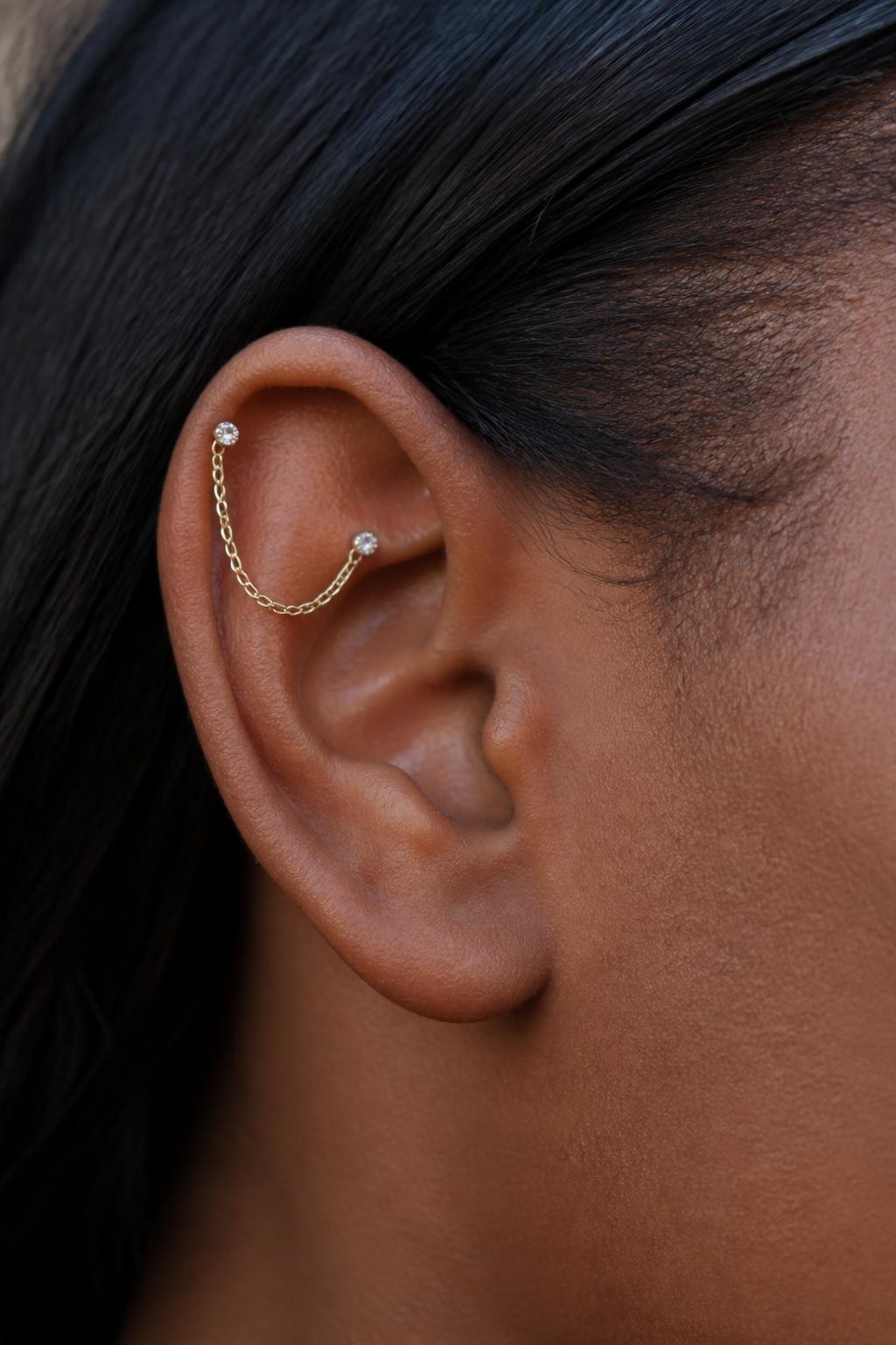Close-up side portrait of a small ear with deep brown skin tone, two petite upper helix piercings connected by a very fine gold chain, tiny crystal studs holding the chain, hair tucked back, soft natural daylight, realistic DSLR detail shot, clean focus on the upper ear and chain.