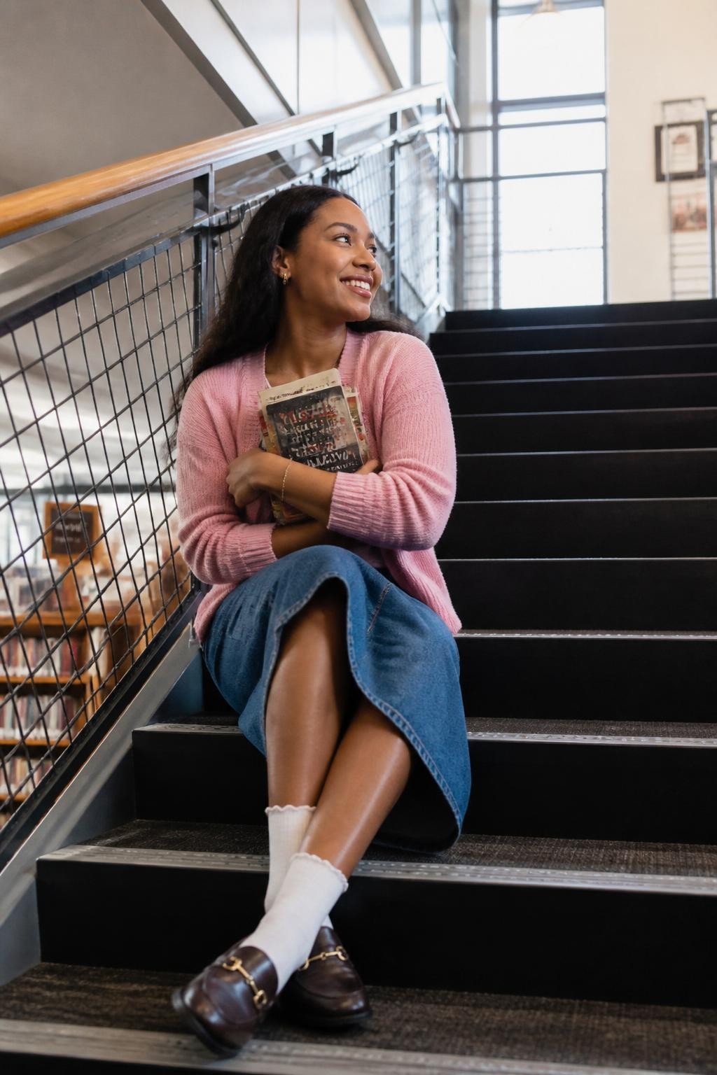 Image prompt: A cute DSLR photo of an adult Black woman with warm brown skin sitting on wide indoor library stairs, wearing a soft pink cardigan, denim midi skirt, white socks, and loafers, hugging two books against her chest while leaning slightly to one side, one knee bent higher than the other, smiling toward the landing instead of directly at the camera, tall windows, metal stair railing, bookshelves visible below, soft daylight, realistic fabric texture, natural skin detail, polished casual library outing photography.