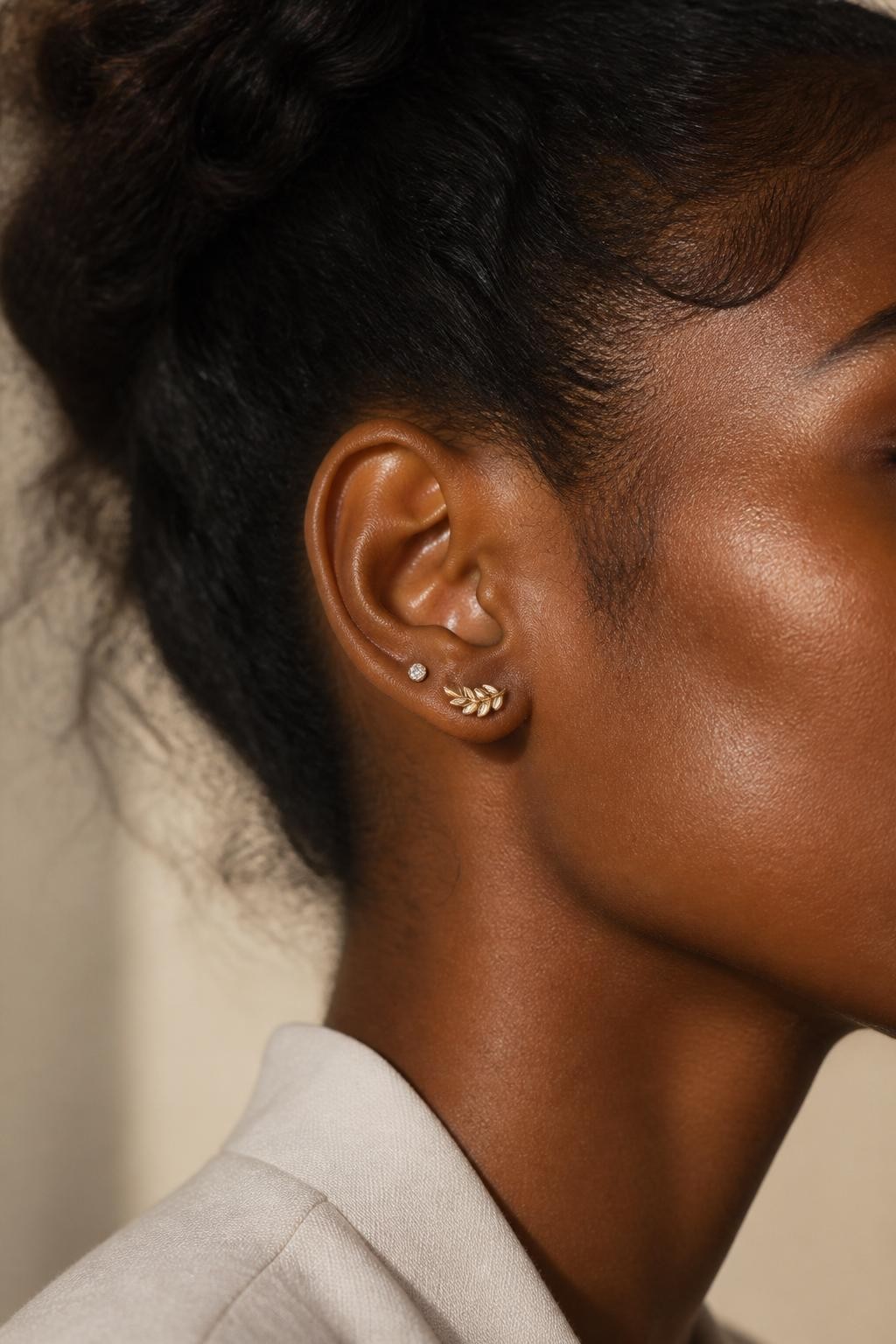 Side-profile beauty portrait of a Black woman’s ear with deep brown skin, soft updo hairstyle, one flat piercing with a curved leaf-like gold stud and one high-lobe piercing with a tiny matching gem, elegant natural light, realistic editorial close-up, refined ear styling.