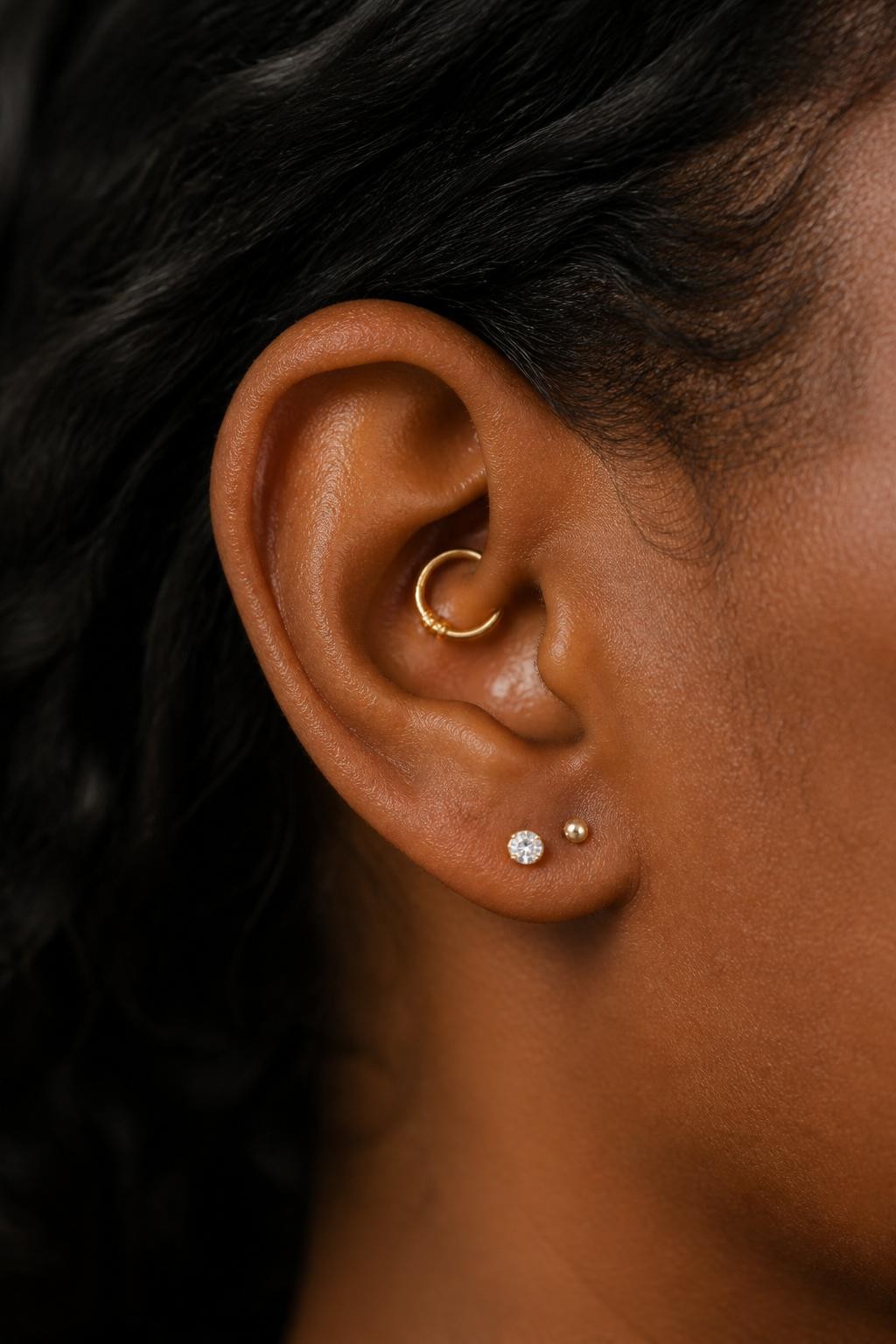 Close-up side view of a small ear with rich brown skin tone, tiny gold daith hoop inside the ear, one small crystal lobe stud, and one petite second lobe dot, hair tucked softly behind the ear, natural light, realistic editorial beauty photography, clear focus on the daith and lobe details.