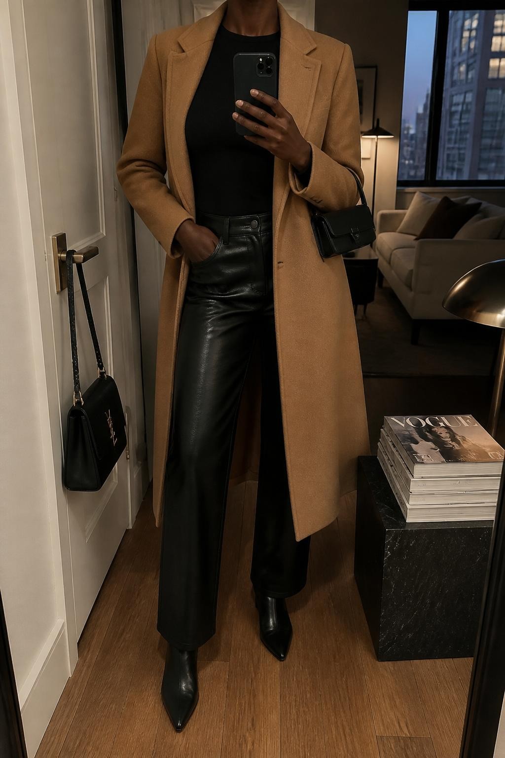 mirror selfie of a Black woman wearing the exact outfit described above, clearly showing a camel long straight wool coat, black fitted long-sleeve top, black faux leather straight-leg pants, black pointed ankle boots, and black mini shoulder bag, holding a phone in front of a mirror, cropped at NECK so the face does not show, with a relaxed casual selfie pose, in a stylish city apartment, warm moody lighting, including a handbag hanging on a door handle and a stack of fashion magazines on a side table, latest iPhone photo quality.