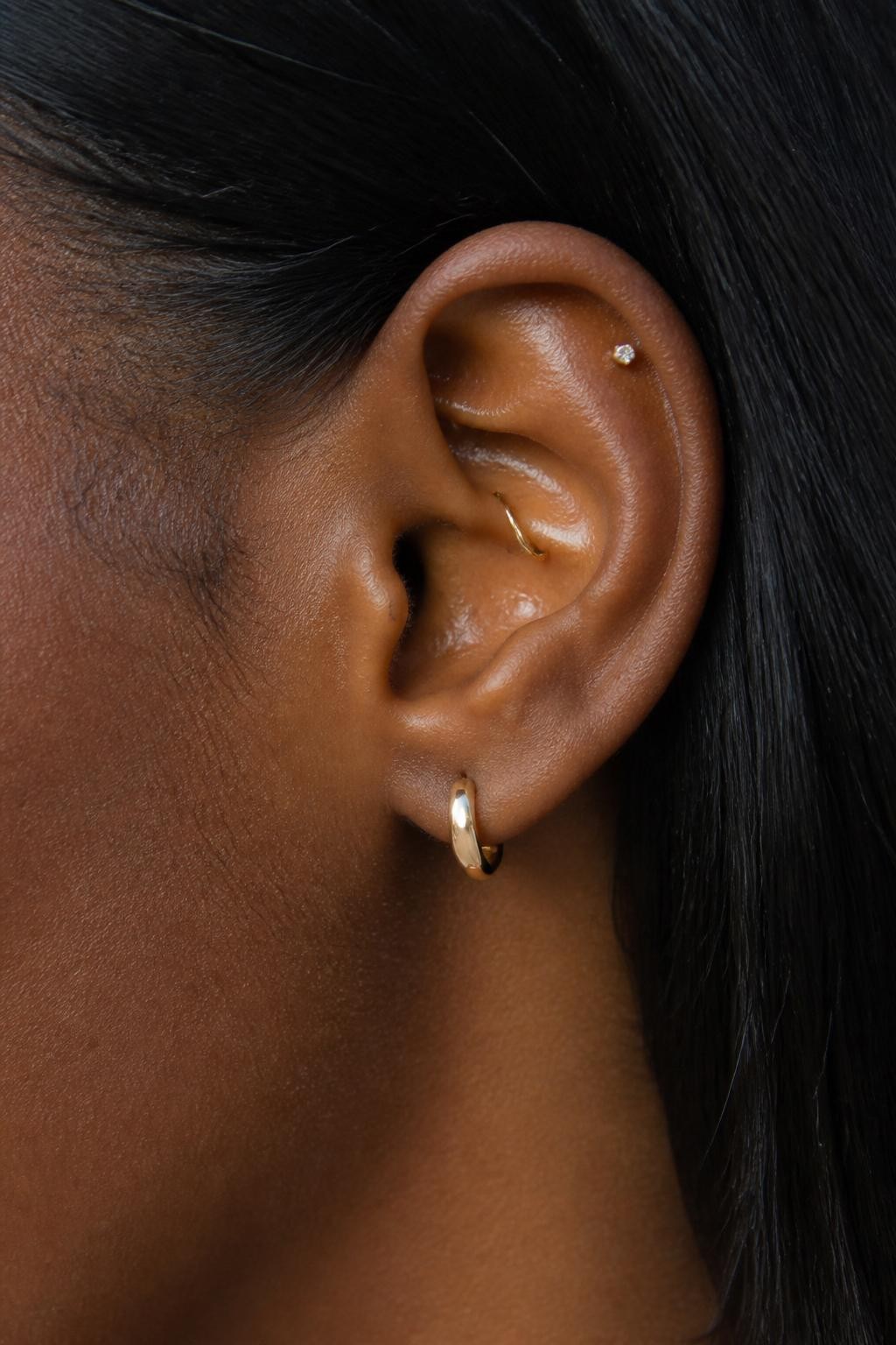Side close-up of an ear with rich dark brown skin tone, one small gold rook hoop, one tiny upper helix stud, and one neat lobe huggie, hair tucked behind the ear, natural daylight, realistic editorial beauty photography, crisp detail on the inner and outer ear jewelry.