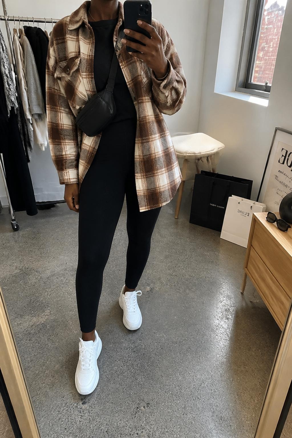 mirror selfie of a Black woman wearing the exact outfit described above, clearly showing a brown and cream plaid oversized shacket, black cotton crew neck tee, black high-waisted thick leggings, white chunky sneakers, and black belt bag worn crossbody, holding a phone in front of a mirror, cropped at NECK so the face does not show, with a relaxed casual selfie pose, in a fashion studio mirror corner, natural window light, including shopping bags beside a chair and sunglasses placed on a dresser, latest iPhone photo quality.