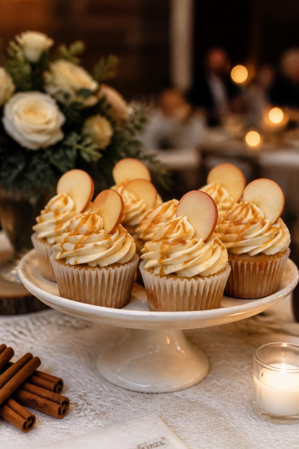 Realistic DSLR close-up of apple cider fall wedding cupcakes with ivory buttercream swirls, thin caramel drizzle, tiny apple slice garnish, cinnamon sticks nearby, cream ceramic cake stand, soft linen tablecloth, warm candlelit reception background.