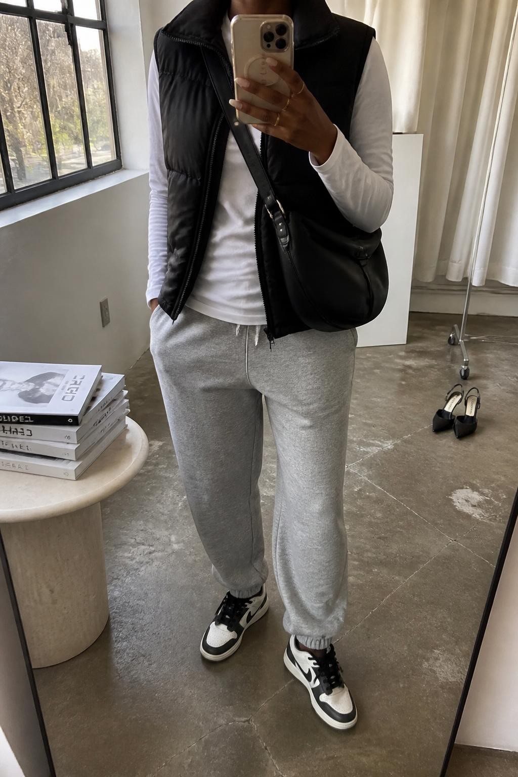 mirror selfie of a Black woman wearing the exact outfit described above, clearly showing a black quilted puffer vest, white long-sleeve cotton tee, grey fleece joggers, black and white sneakers, and black nylon crossbody bag, holding a phone in front of a mirror, cropped at NECK so the face does not show, with a relaxed casual selfie pose, in a fashion studio mirror corner, natural window light, including a pair of heels on the floor nearby and stack of fashion magazines on a side table, latest iPhone photo quality.
