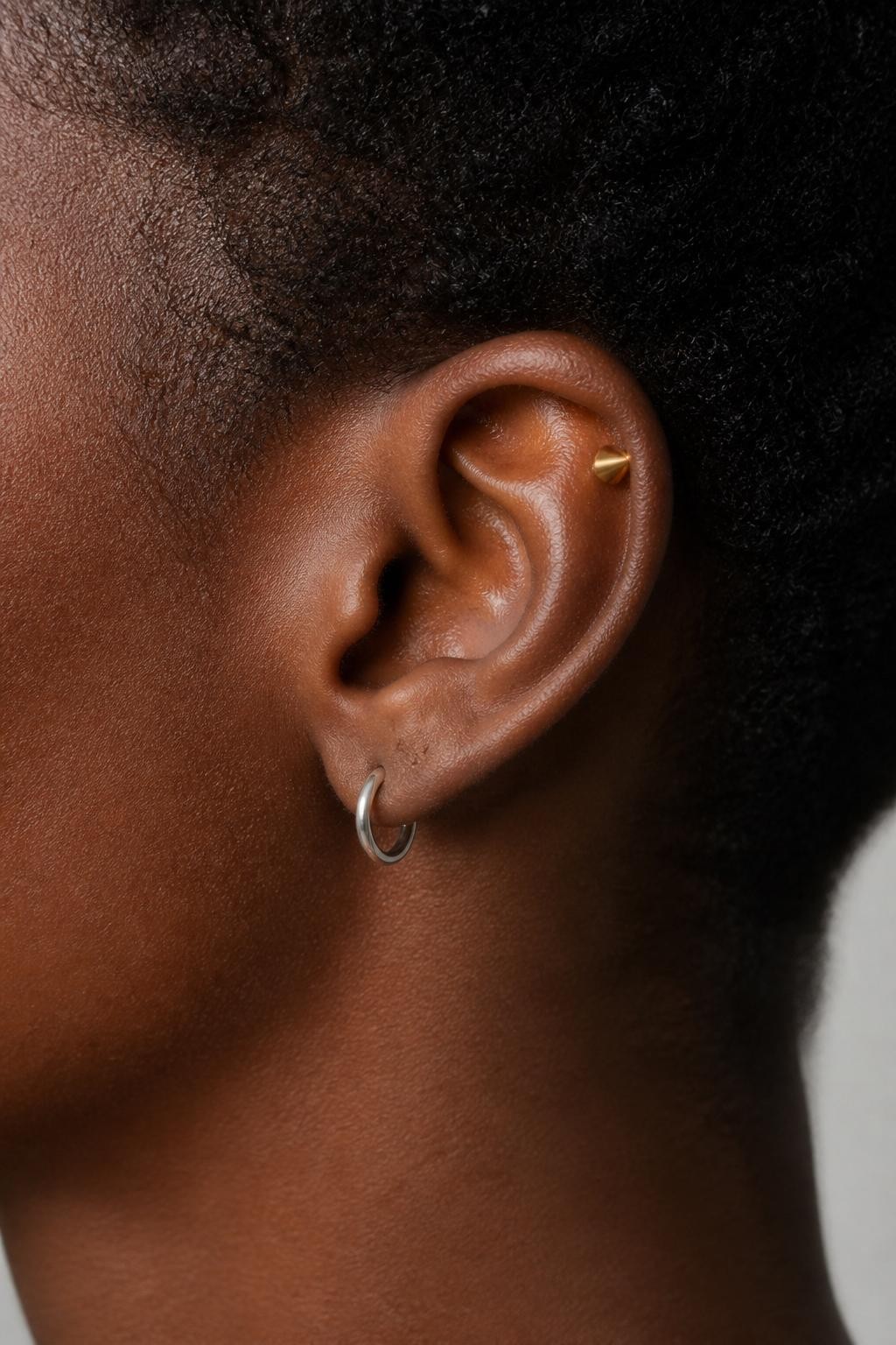 Detailed side ear close-up of a Black woman with deep brown skin and short natural hair tucked behind the ear, one snug piercing with a slim silver hoop and one helix piercing with a small gold spike stud, high-end beauty photography, soft light, sharp focus on the ear and jewelry.