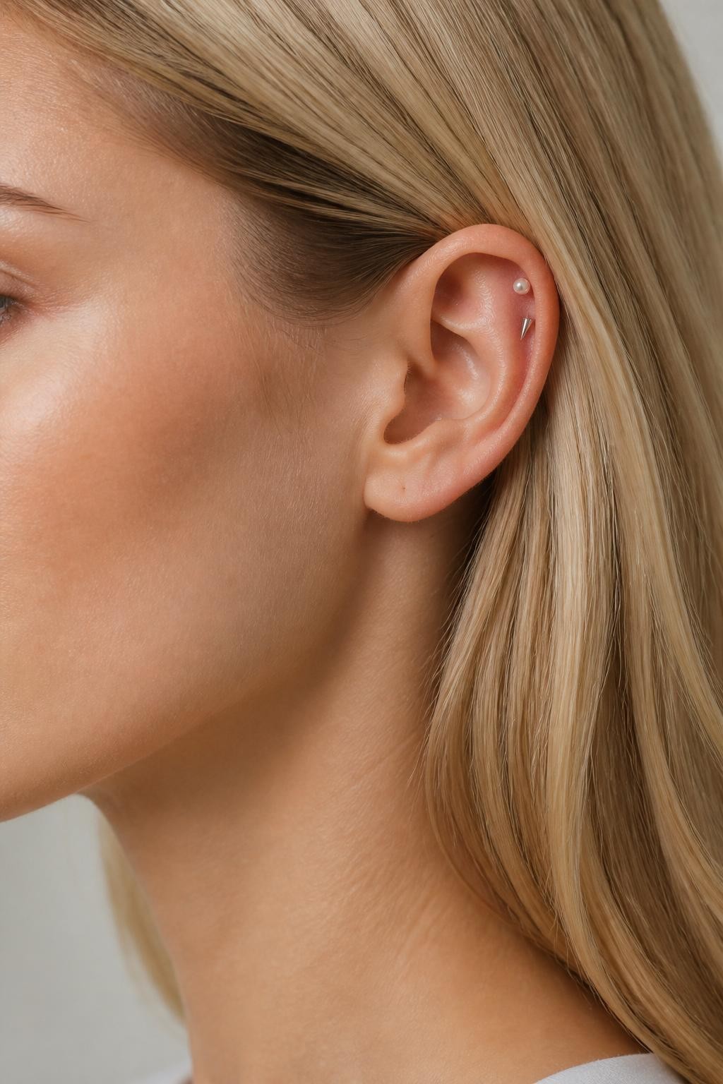 Detailed side ear portrait of a woman with light neutral skin tone, blonde hair tucked behind the ear, two helix piercings styled with one tiny pearl stud and one slim silver spike, soft editorial beauty light, crisp ear detail, clean minimal background.