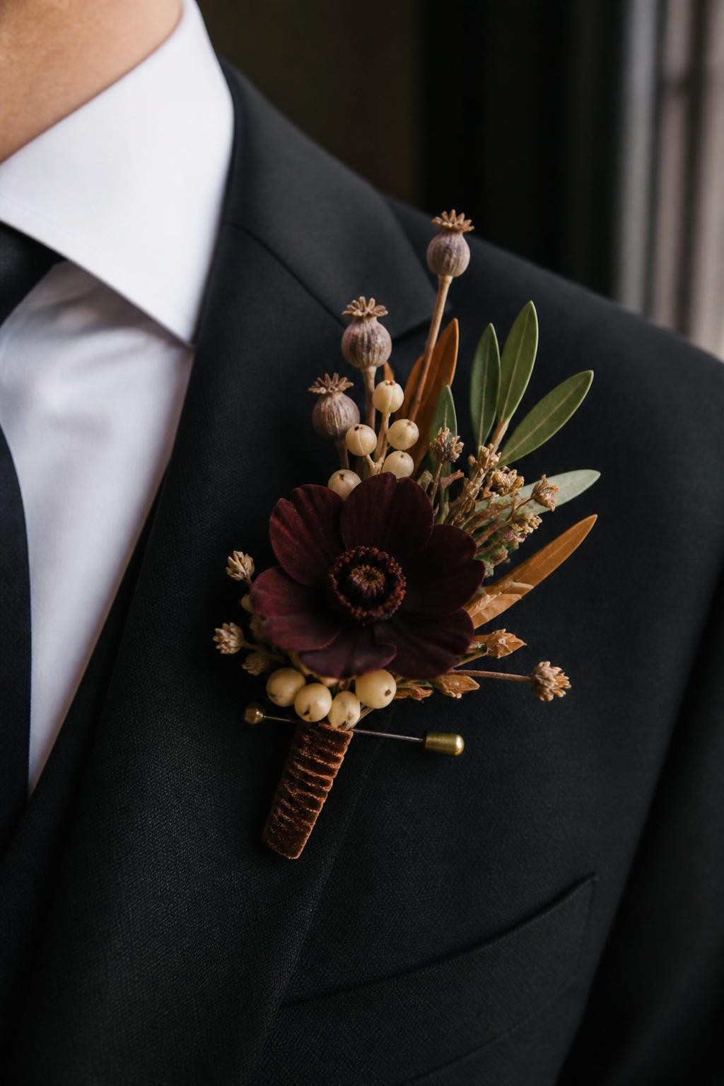 Realistic DSLR macro photo of a fall wedding boutonniere pinned to a black suit lapel, chocolate cosmos bloom, tiny cream berries, dried seed pods, olive leaves, brown velvet ribbon wrap, brass boutonniere pin, soft window light, groom face cropped out, elegant autumn wedding detail.