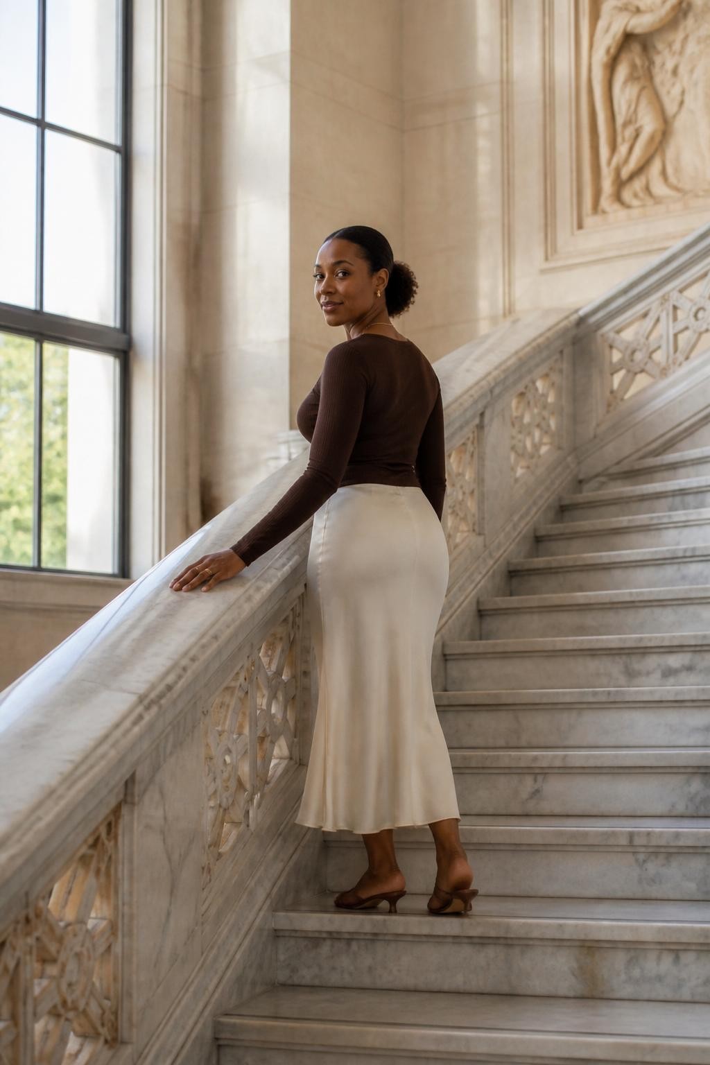 Image prompt: A beautiful DSLR museum photo of an adult Black woman with warm brown skin standing halfway up a wide marble staircase, wearing a fitted chocolate brown long-sleeve top, ivory slip skirt, and low heels, one hand lightly grazing the stone railing while her body turns back toward the camera, face calm with a small smile, staircase lines leading upward behind her, tall museum windows, soft afternoon light, realistic marble veining, natural hair pulled back neatly, subtle jewelry, elegant travel outfit, modern museum architecture photography with a human feel.