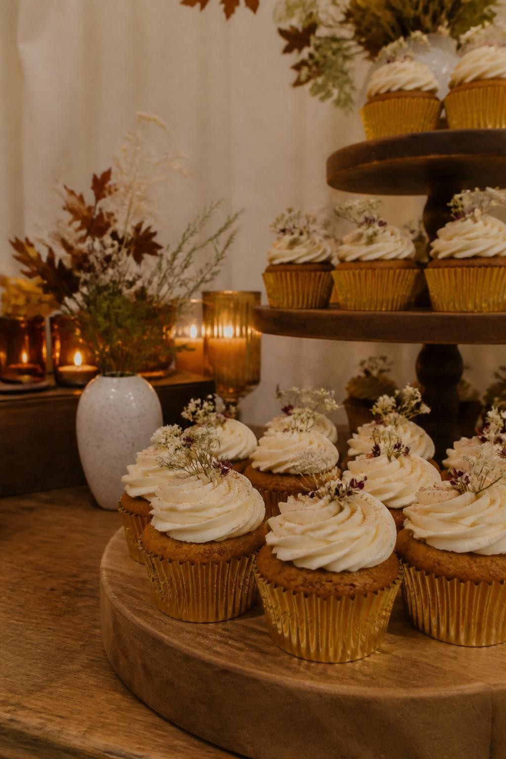 Realistic DSLR photo of fall wedding cupcakes on a wooden dessert table, brown butter pumpkin cupcakes in gold liners, cream cheese frosting piped into soft rosettes, tiny cinnamon dusting, small dried flowers, amber candles, linen backdrop, warm indoor wedding lighting, no people visible.
