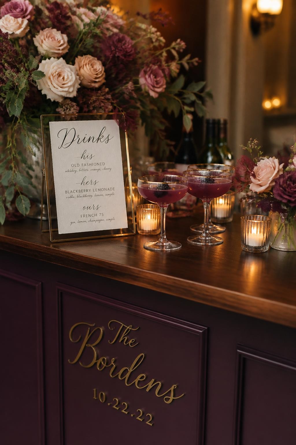 Realistic DSLR photo of a fall wedding cocktail bar with plum painted front, dusty pink floral arrangements, purple signature cocktails in coupe glasses, gold-rimmed glassware, small candle clusters, handwritten drink menu, dark wood counter, warm moody reception lighting.