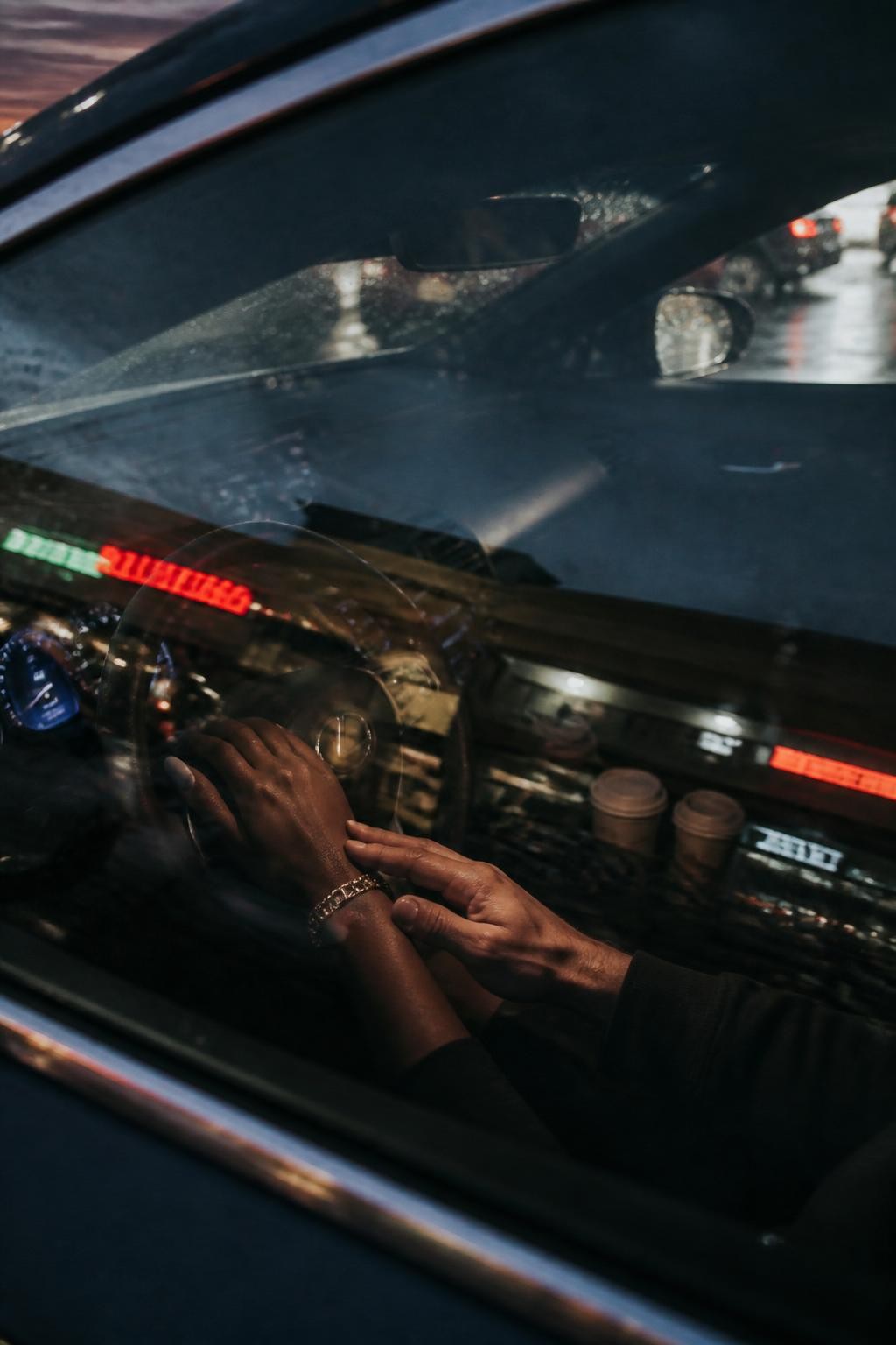 Image prompt: A modern no-face couple photo captured through a car window at dusk, featuring an adult couple sitting in the front seats of a parked car, their faces fully obscured by soft window reflection and intentional blur, a Black woman with deep brown skin visible through her hand resting on the steering wheel and a gold bracelet, her partner’s hand gently placed over hers, dashboard lights glowing, raindrops on the glass, blurred neon signs reflected across the window, takeaway coffee cups in the cup holders, realistic glass glare, cinematic DSLR couple photography, private and stylish.