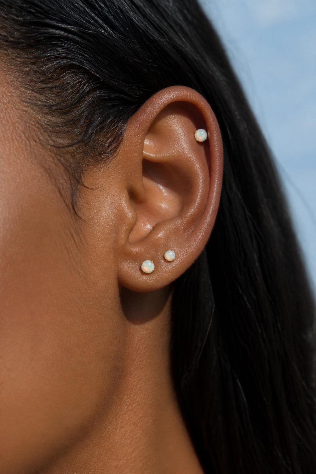 Close-up side portrait of a small ear with rich dark brown skin tone, three tiny opal studs scattered lightly across the lobe and upper cartilage, soft color-shifting stones, hair pulled behind the ear, natural daylight, realistic DSLR editorial image, clear focus on the delicate opal jewelry.