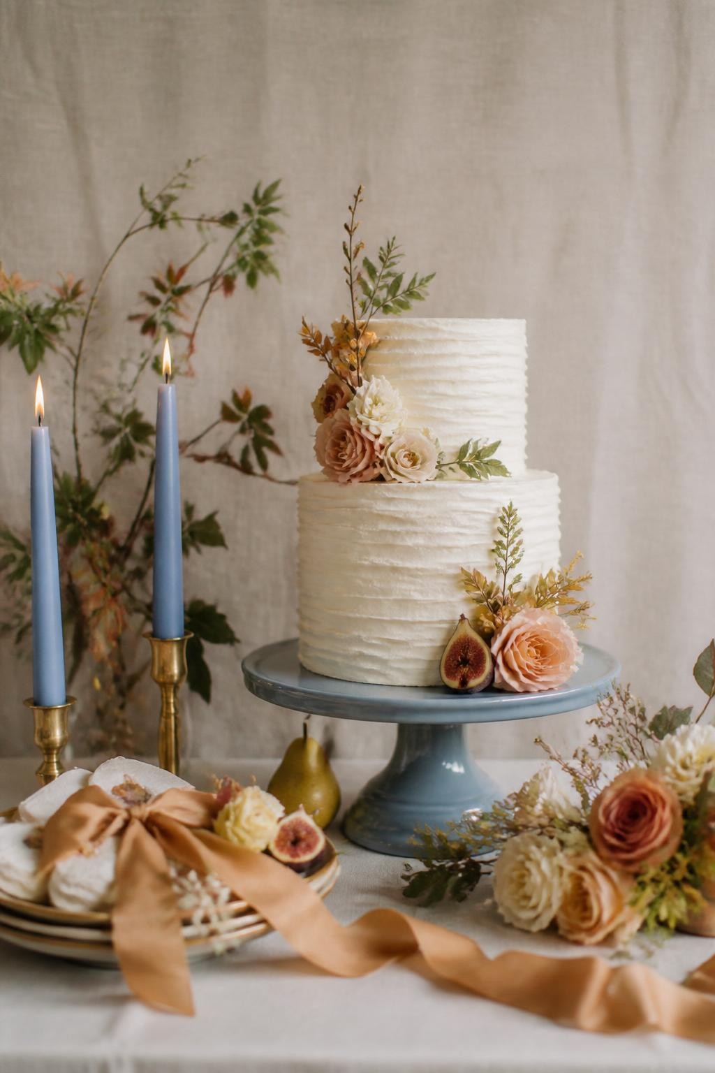 Realistic DSLR photo of a fall wedding dessert table with butterscotch silk ribbons, French blue cake stand, toasted almond linen backdrop, cream buttercream wedding cake, small blue taper candles, pears, figs, and delicate fall flowers, soft natural light, editorial detail shot.