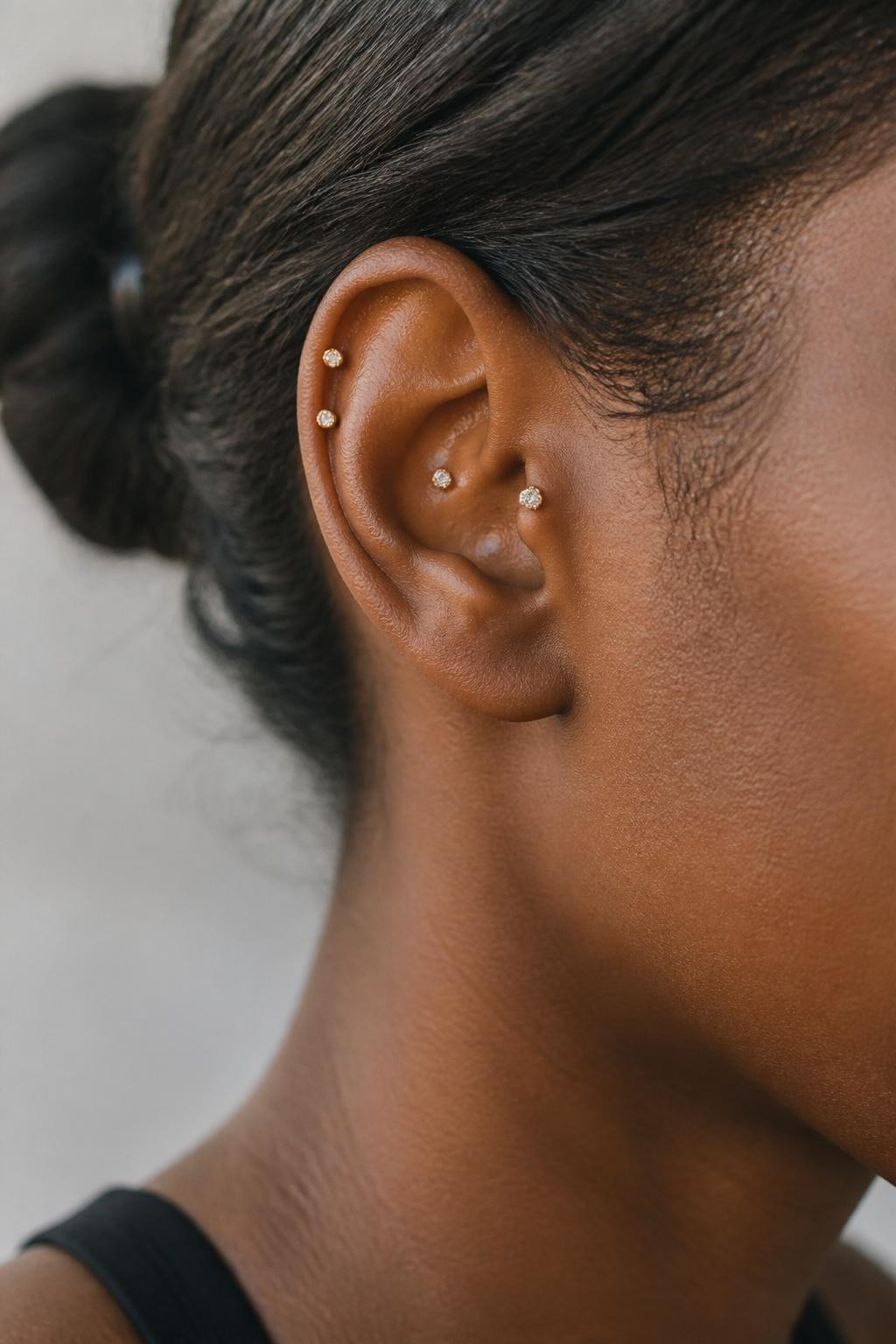 Side portrait close-up of an ear with warm deep brown skin tone, two tiny helix piercings along the outer ear and one small crystal stud on the flat cartilage, hair tied back, soft natural daylight, realistic editorial beauty image, focus on upper ear placements and fine jewelry.
