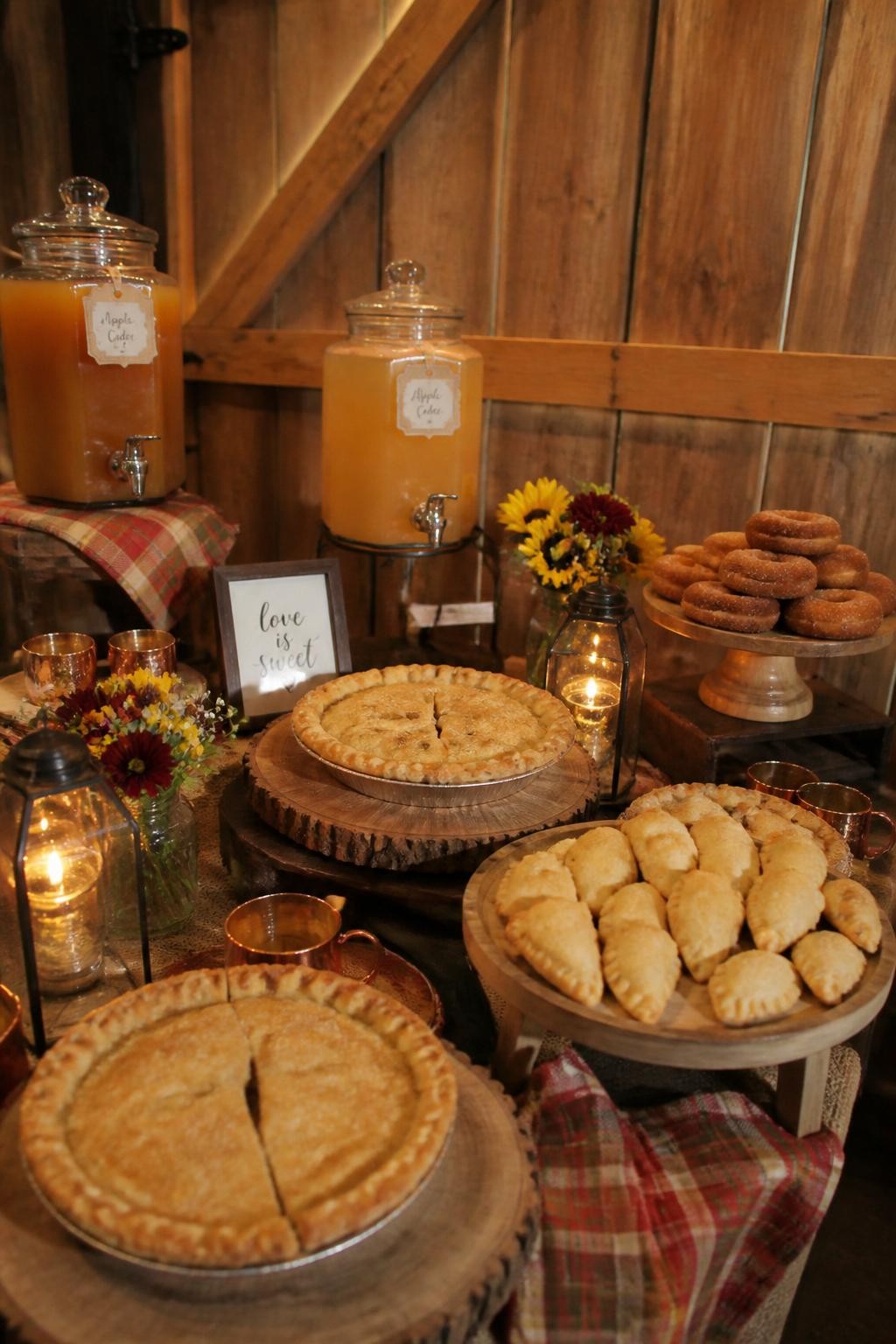 Realistic DSLR photo of a rustic fall wedding dessert table with apple pies, mini hand pies, cider donuts, glass cider dispensers, wooden cake stands, plaid napkins, copper mugs, small fall flowers, candle lanterns, barn wall background, warm cozy lighting.