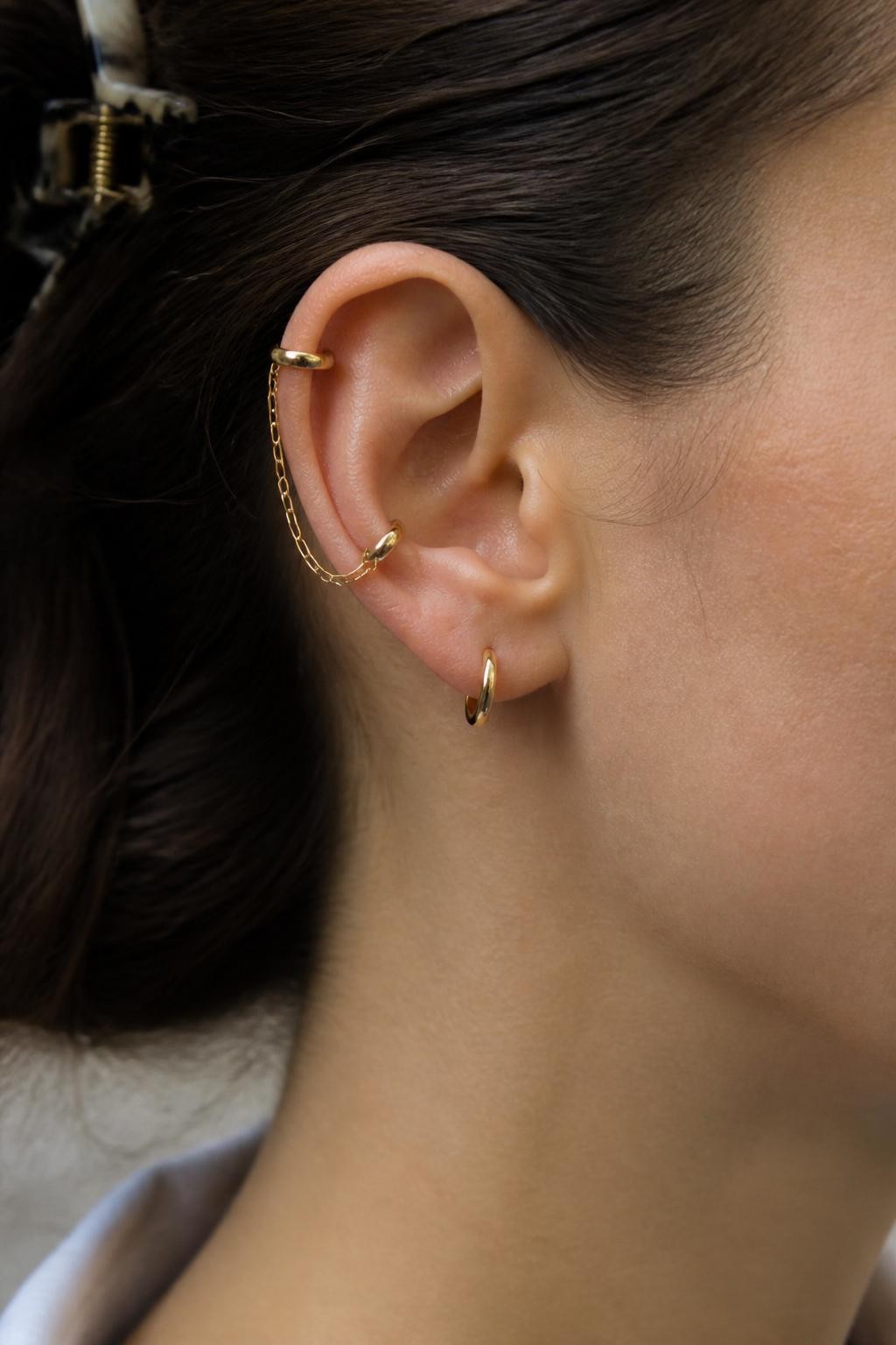 Side portrait close-up of an ear with neutral light skin tone, two upper helix piercings connected by a fine gold chain and one small lobe huggie below, hair clipped behind the ear, soft daylight, realistic DSLR editorial shot, sharp focus on the chain detail and ear stack.