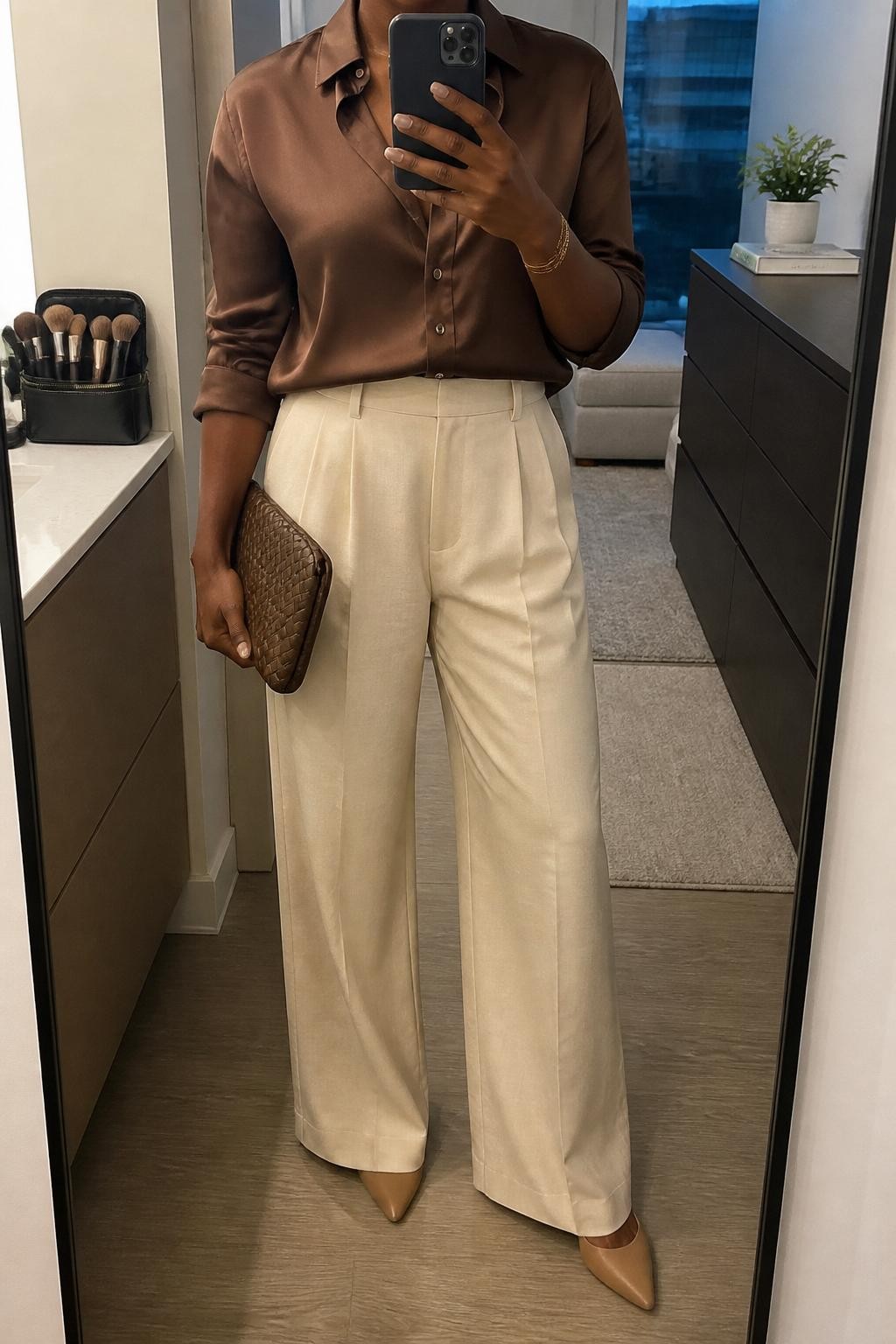 mirror selfie of a Black woman wearing the exact outfit described above, clearly showing a chocolate brown satin button-up blouse, cream high-waisted wide-leg trousers, nude pointed flats, brown woven clutch, and gold layered bracelet, holding a phone in front of a mirror, cropped at NECK so the face does not show, with a relaxed casual selfie pose, in a stylish city apartment, soft indoor lighting, including a makeup brush or cosmetic bag on the counter and a small plant on a dresser, latest iPhone photo quality.