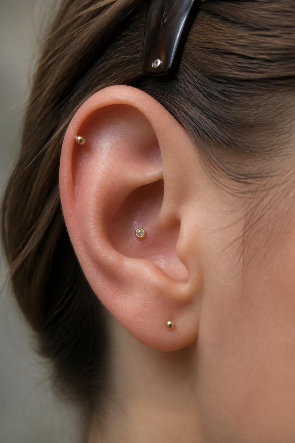 Close-up side view of a small ear with light neutral skin tone, one tiny conch stud in the inner ear and one small gold dot on the upper helix, minimal lobe stud below, hair clipped back, soft natural light, realistic editorial beauty shot, focus on the inner and upper ear details.
