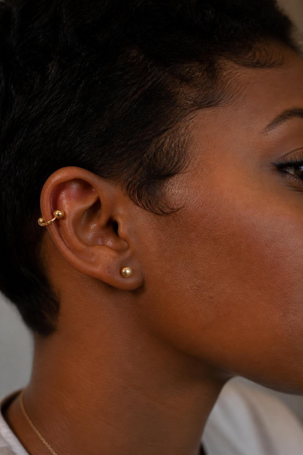 Detailed side ear portrait of a Black woman with warm espresso-toned skin and short hair tucked behind the ear, two helix piercings on the upper outer ear connected by a single sleek gold orbital-style ring, modern beauty photography, soft natural light, crisp detail on cartilage jewelry, minimal neutral background.