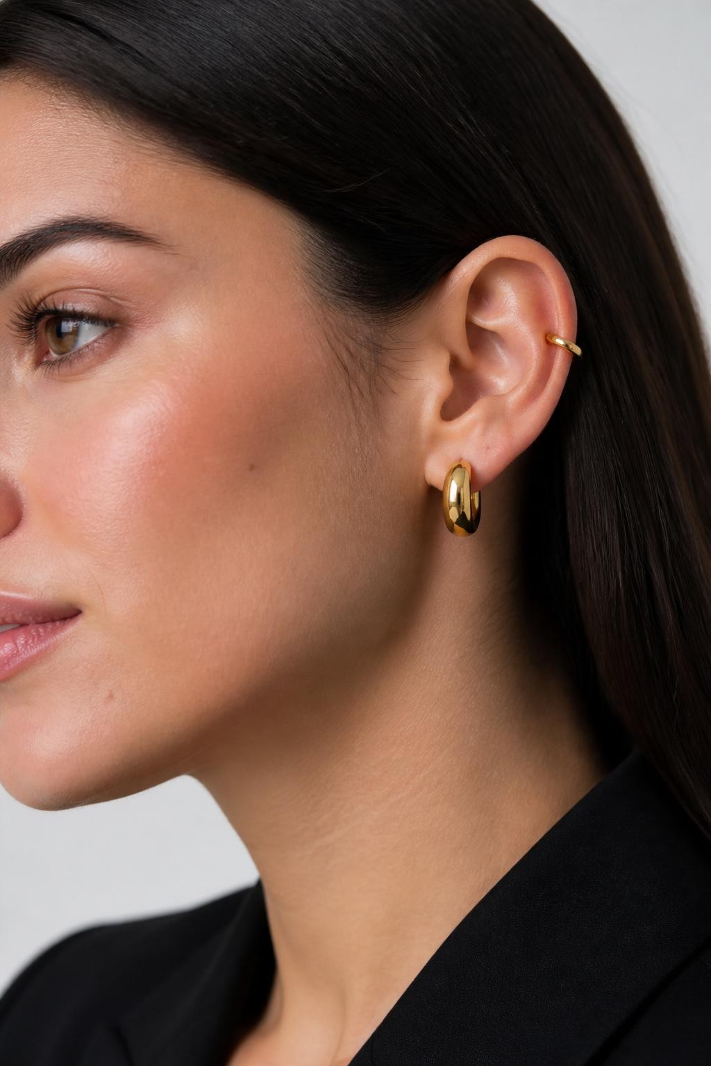 Close-up side portrait of a woman with neutral light skin tone, dark hair tucked neatly behind the ear, one crescent-shaped rook ring and one chunky lower-lobe huggie in polished gold, soft clean studio lighting, realistic beauty photography, sharp focus on the ear and jewelry.