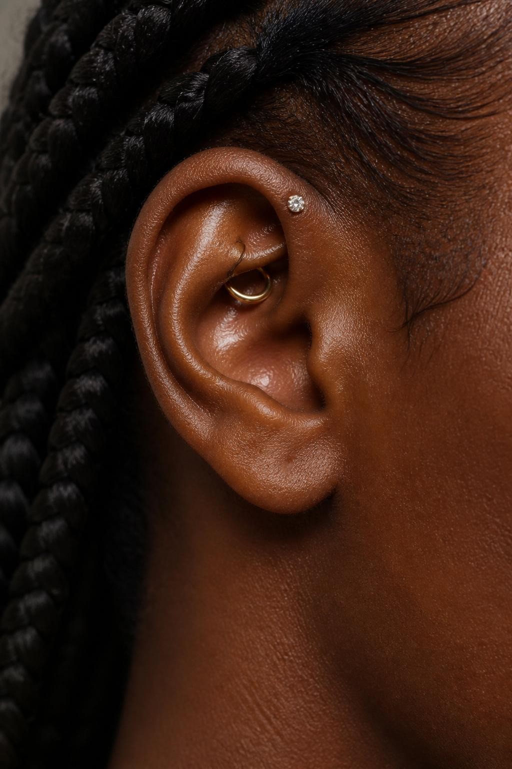 Close-up portrait of an ear with deep brown skin tone, small gold hoop through the rook, single floating crystal stud on the flat upper cartilage, hair braided back away from the ear, smooth natural light, realistic DSLR beauty shot, focus on inner upper ear placement and jewelry shine.
