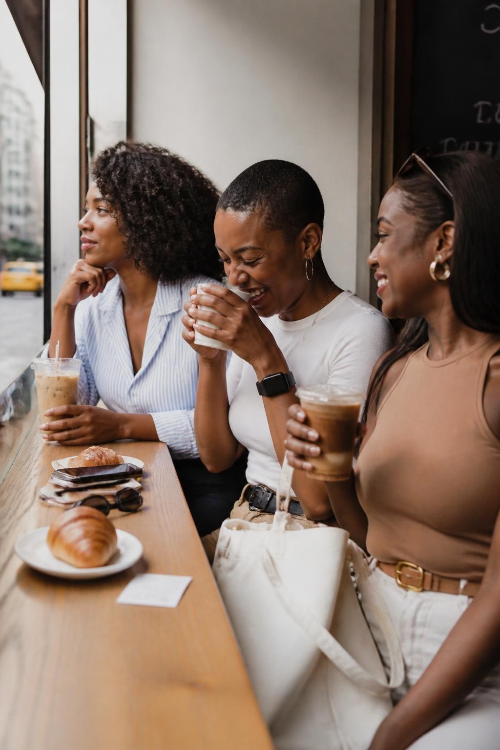 Image prompt: A realistic DSLR cafe photo of three adult Black women friends sitting side by side at a window counter, wearing casual stylish outfits in soft blue, white, and tan, each holding a different cafe drink, one looking out the window, one laughing into her cup, one turning toward the others with a smile, croissants, phone, sunglasses, tote bag, and small receipt on the counter, city street blurred outside, soft morning light on their skin, realistic coffee condensation, natural hair textures, clean modern cafe mood, candid friendship lifestyle photography.