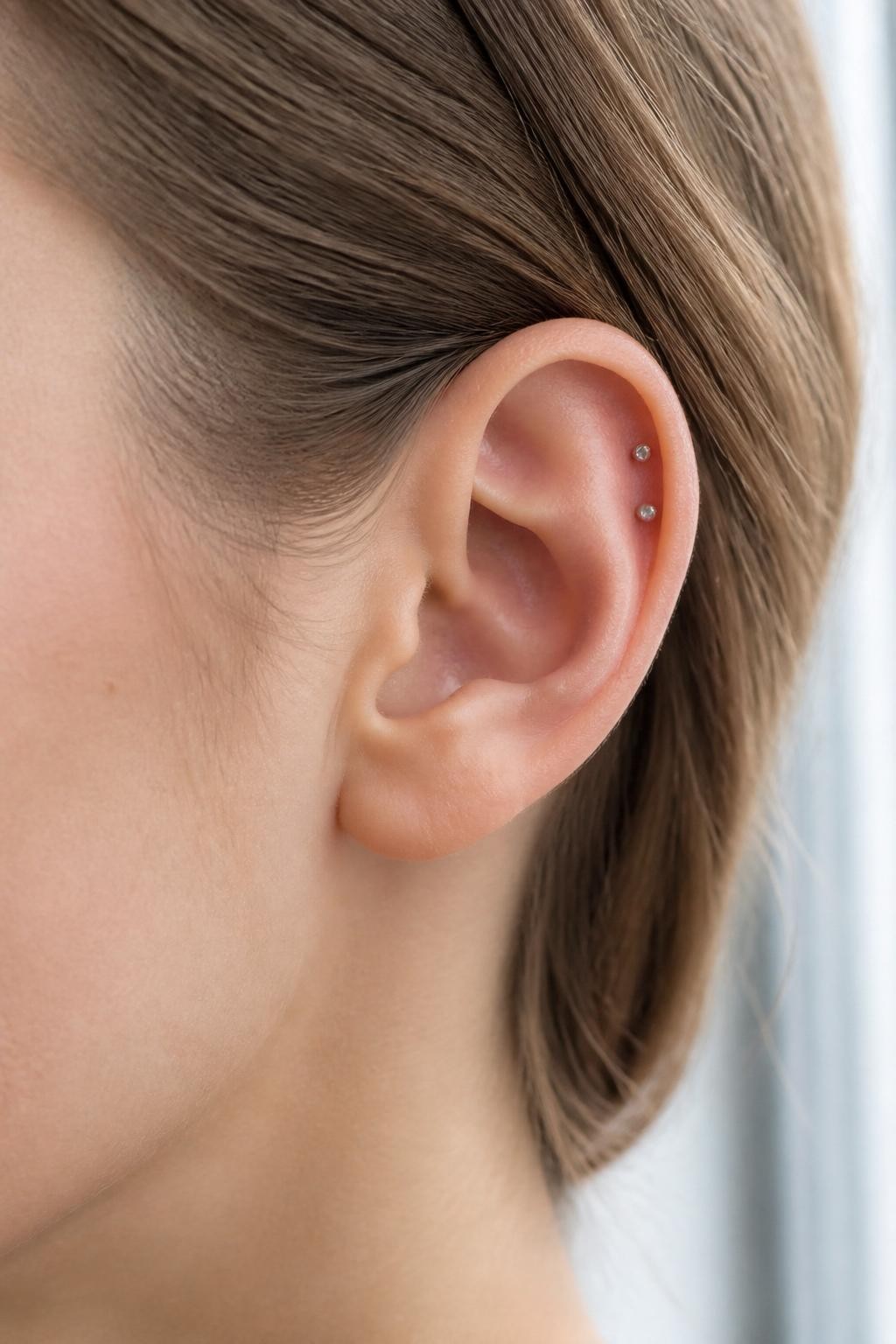 Side portrait close-up of a small ear with neutral fair skin tone, two tiny silver studs placed on the forward helix near the front curve of the ear, sleek tucked hair, soft window light, realistic beauty editorial style, sharp focus on the forward helix area.
