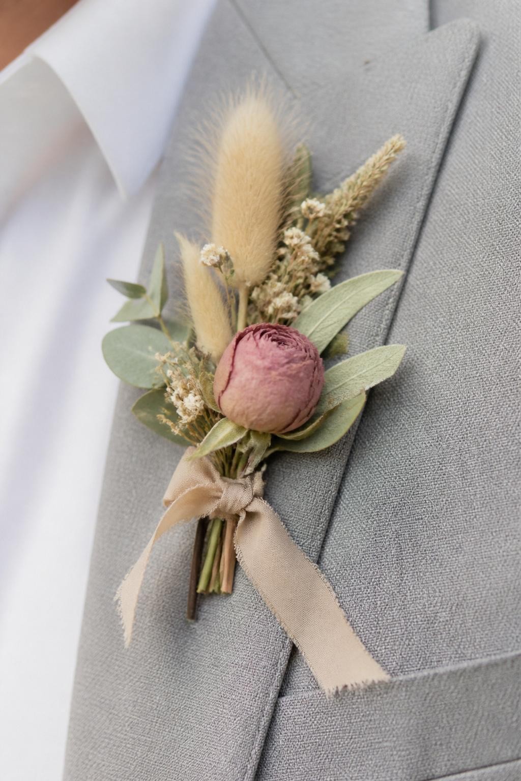 Realistic DSLR close-up of a soft fall wedding boutonniere with dried bunny tail grass, mauve rosebud, tiny cream flowers, dusty green leaves, beige silk ribbon, pinned to a light gray suit lapel, soft natural daylight, romantic minimalist wedding detail.