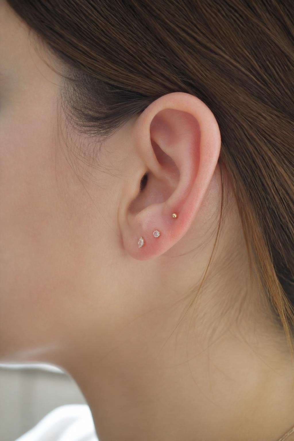 Side portrait close-up of a small ear with neutral fair skin tone, three tiny studs fanning from the upper lobe toward the lower cartilage, one gold bead, one crystal, one tiny marquise stud, hair tucked behind ear, soft daylight, realistic DSLR beauty shot, focus on the compact fan arrangement.