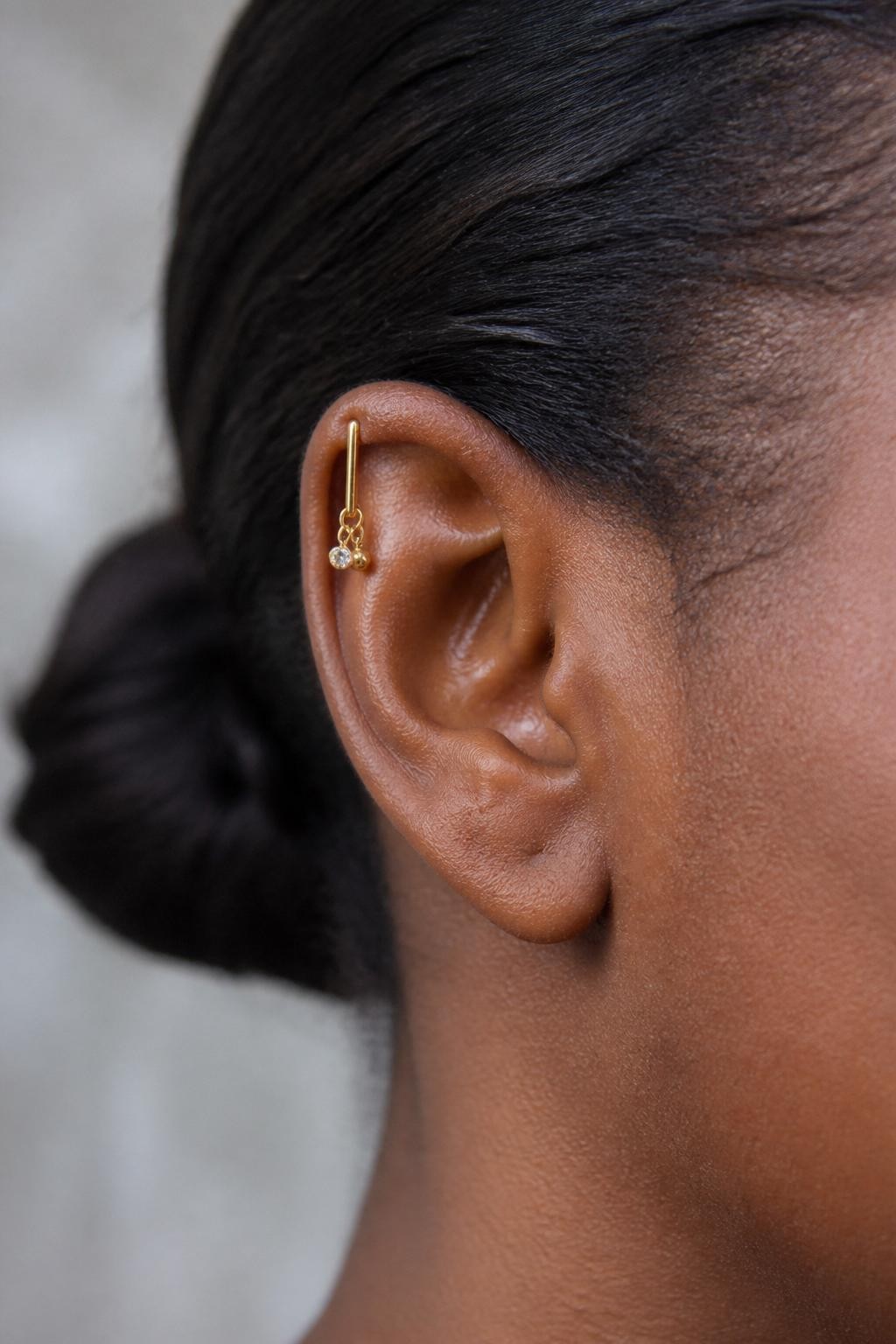 Side portrait close-up of an ear with rich brown skin tone, vertical upper helix piercing styled with a slim gold bar and two tiny dangling charms, one mini crystal charm and one tiny bead charm, hair pulled into a low bun, natural light, realistic DSLR detail shot, clean focus on upper ear.