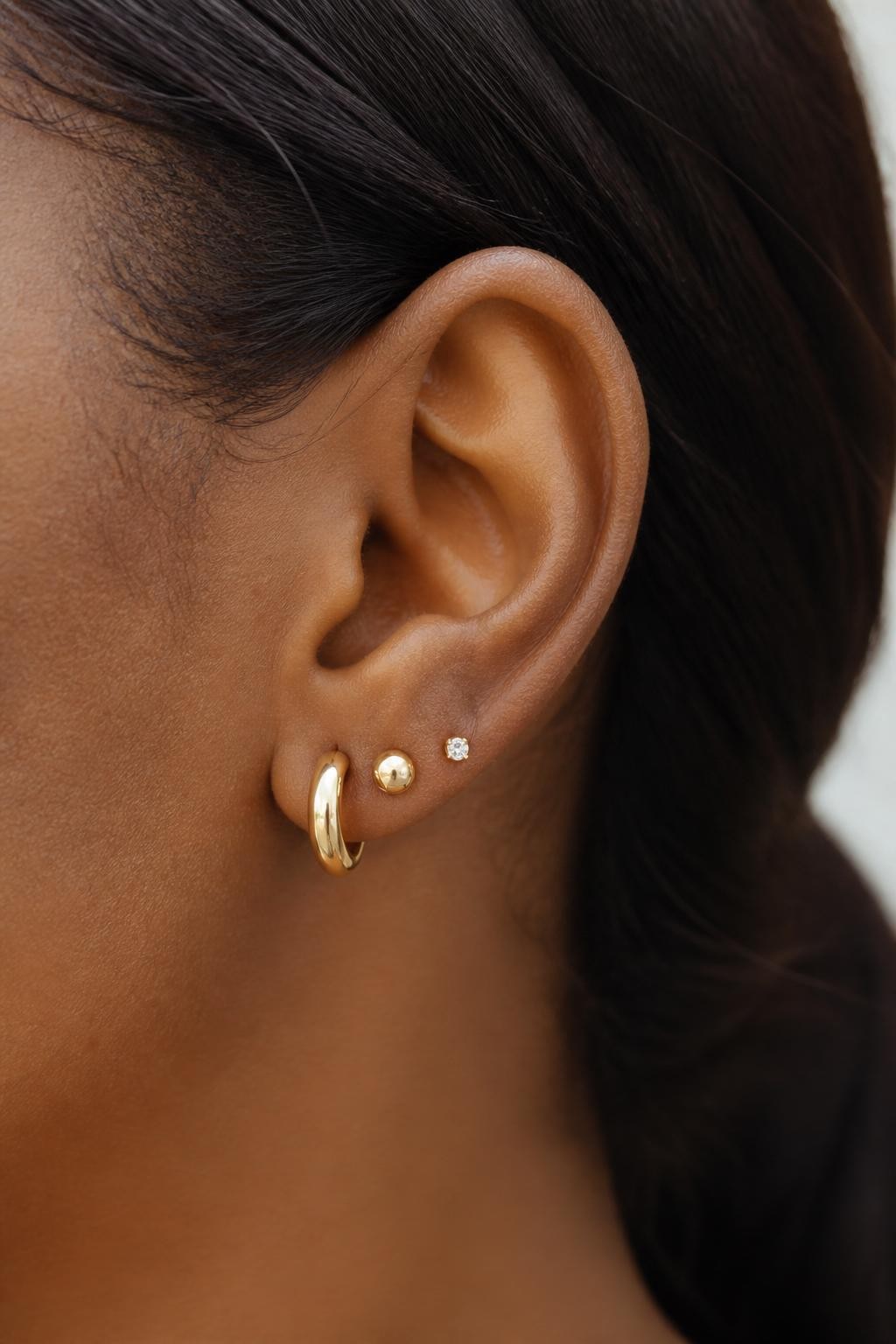 Close-up side portrait of an ear with dark brown skin tone, three lobe piercings styled with a size gradient of gold jewelry from largest huggie to medium stud to tiny crystal, hair softly tucked back, clean soft background, realistic DSLR photo, sharp focus on the lobe area.