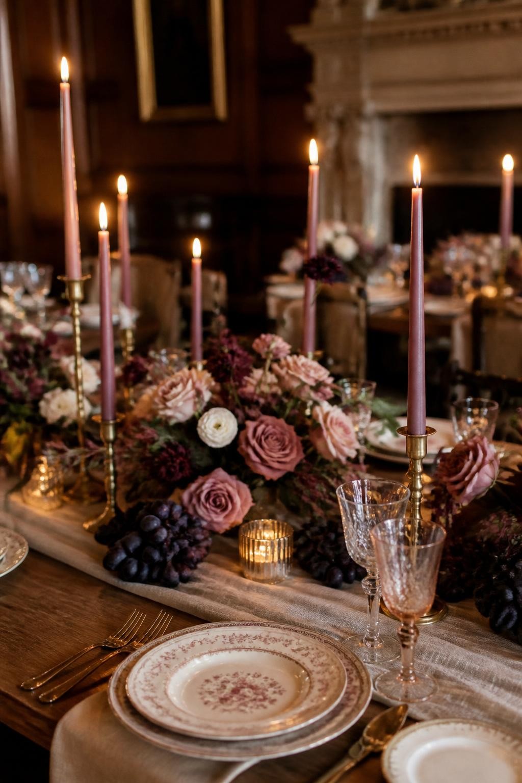 Realistic DSLR photo of a fall wedding tablescape with blush pink taper candles in brass holders, low plum floral centerpieces, mauve roses, cream lisianthus, dark purple grapes, linen runner, vintage china plates, warm candlelit manor reception, no visible faces.