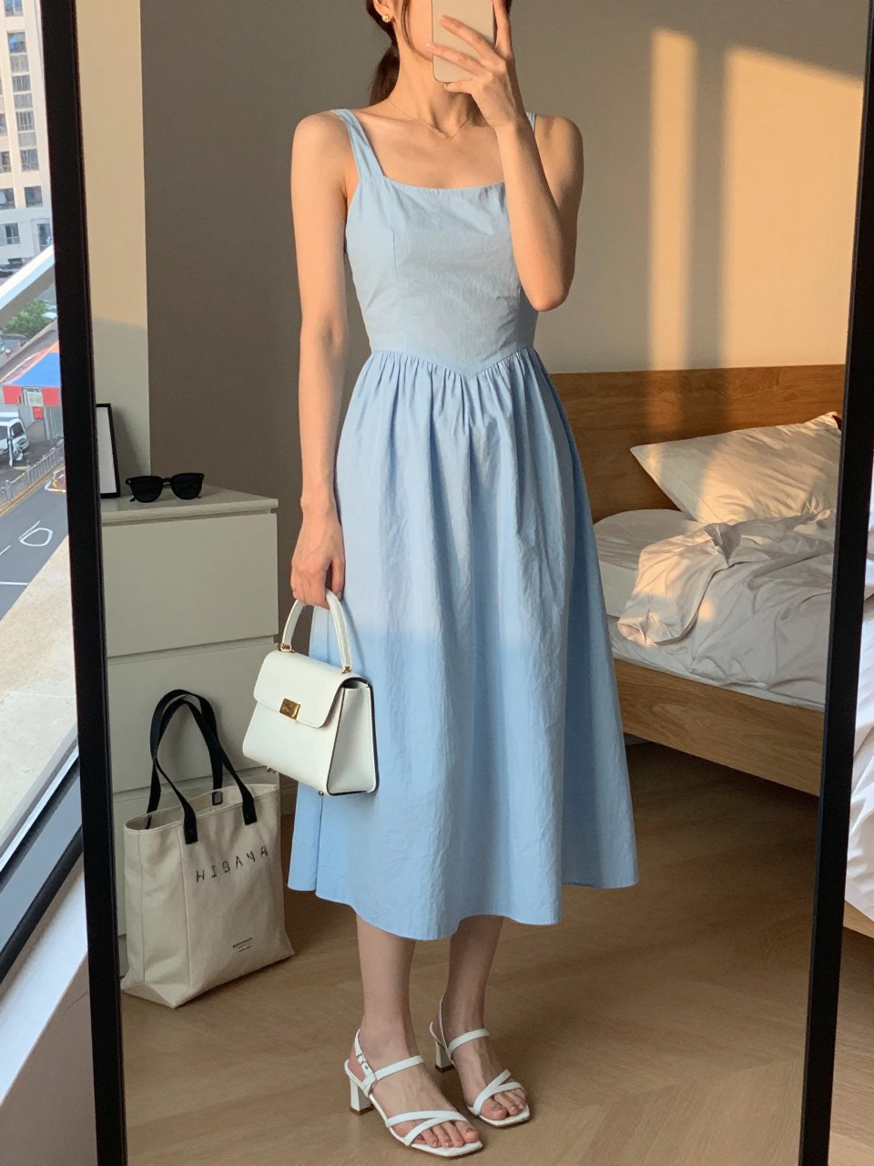 Image prompt mirror selfie of a woman wearing a soft sky blue cotton midi sundress with white slim-strap heeled sandals, a white mini handbag, simple gold stud earrings, and hair in a sleek ponytail, holding a phone in front of a mirror, cropped at NECK so face doesn't show, with a natural casual selfie pose, in a city apartment, evening golden hour, including small lived-in details like a tote bag on the floor, sunglasses on a dresser, or soft bedding in the background, latest iPhone photo quality.