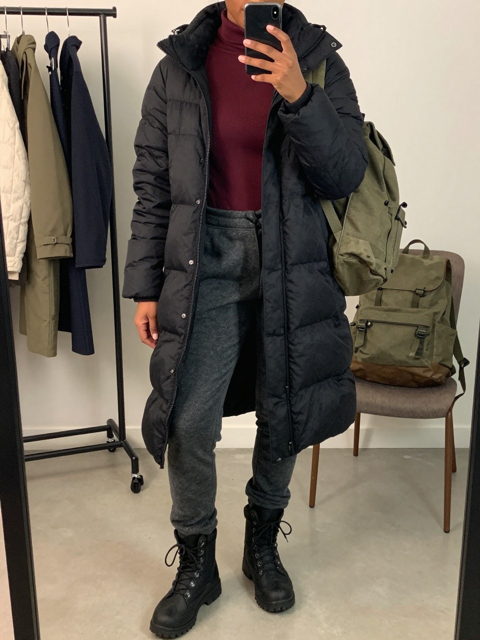 Mirror selfie of a Black woman wearing a black thigh-length insulated puffer coat over a fitted burgundy thermal mock-neck top, charcoal-grey fleece-lined hiking trousers, black waterproof lace-up boots, and an olive-green canvas backpack, clearly showing the exact clothing items, colors, fabrics, and accessories listed, holding a phone in front of a fashion studio mirror corner, cropped at the neck so the face does not show, relaxed casual selfie pose, soft indoor lighting, a clothing rack with garments behind and a backpack resting beside a chair, natural outdoor-ready setting, latest iPhone photo quality.