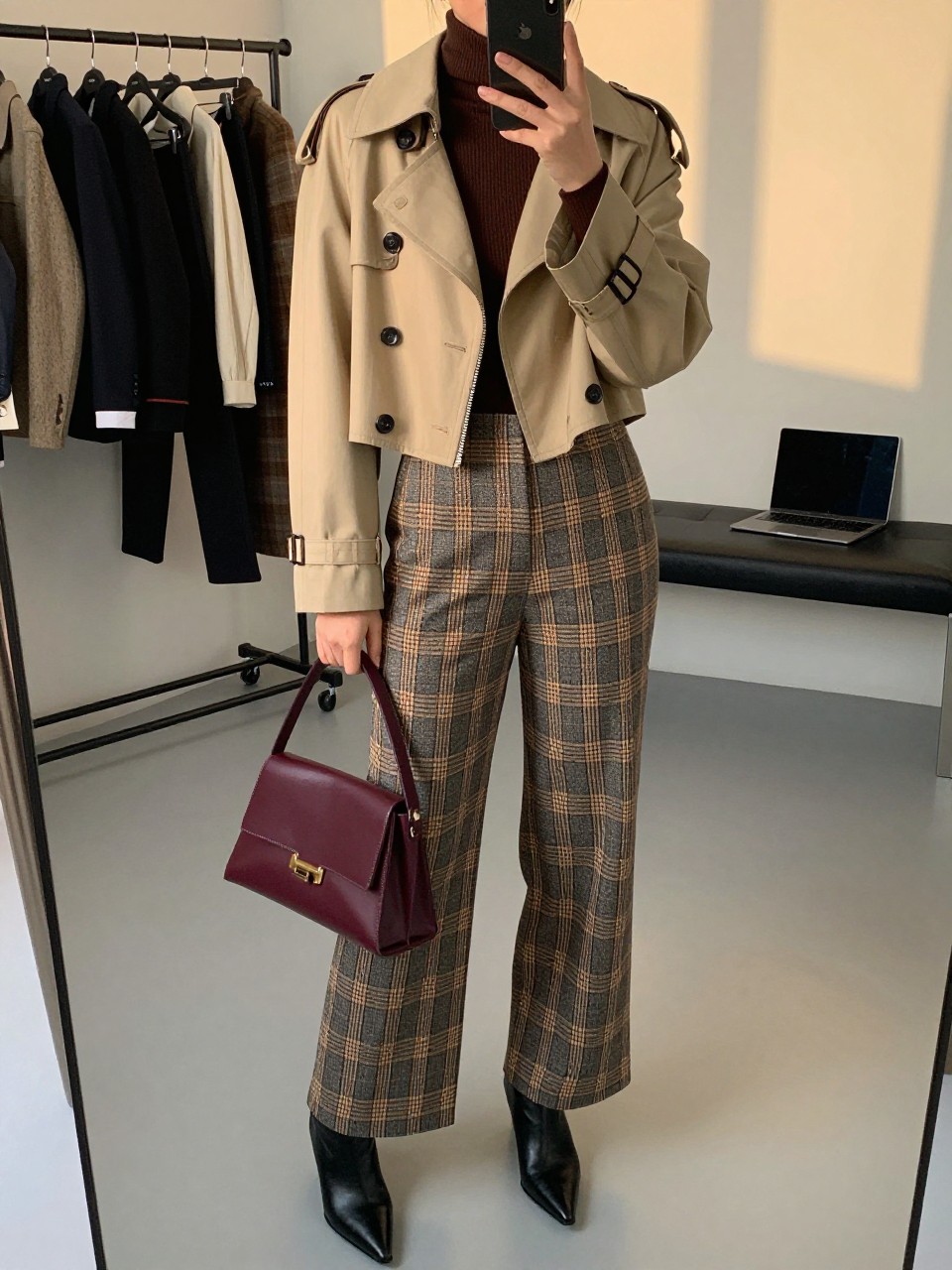 Mirror selfie of a woman wearing a cropped beige trench jacket, fitted chocolate-brown turtleneck, charcoal and camel plaid wide-leg trousers, black pointed-toe ankle boots, and a structured burgundy handbag, clearly showing the exact clothing items, colors, fabrics, and accessories listed, holding a phone in front of a fashion studio mirror corner, cropped at the neck so the face does not show, relaxed casual selfie pose, evening golden-hour lighting, a clothing rack with garments behind and a laptop on a bench, polished realistic setting, latest iPhone photo quality.