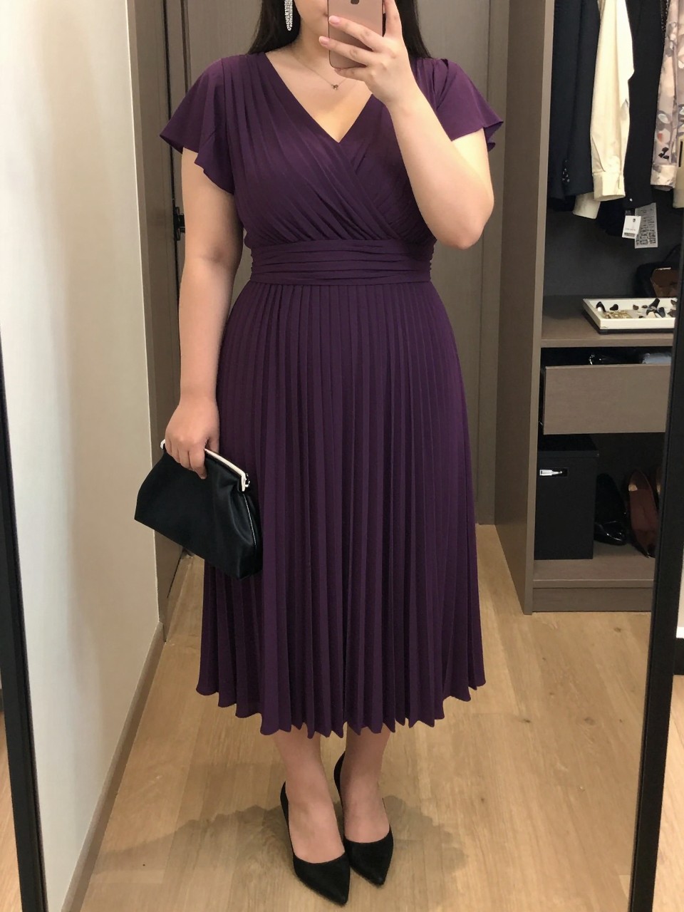 Mirror selfie of a plus size woman wearing a plum purple pleated midi dress with short flutter sleeves, a modest V-neckline, and a softly fitted waist, black pointed kitten heels, a small black satin clutch, and silver crystal drop earrings, clearly showing the exact dress, plum pleats, black footwear, clutch, and accessories listed, holding a phone in front of a mirror, cropped at the neck so the face is hidden, relaxed graceful selfie pose, in a walk-in closet mirror with soft indoor lighting, an open closet with clothes visible and a jewelry tray on the vanity, realistic romantic dinner-date outfit, latest iPhone photo quality.