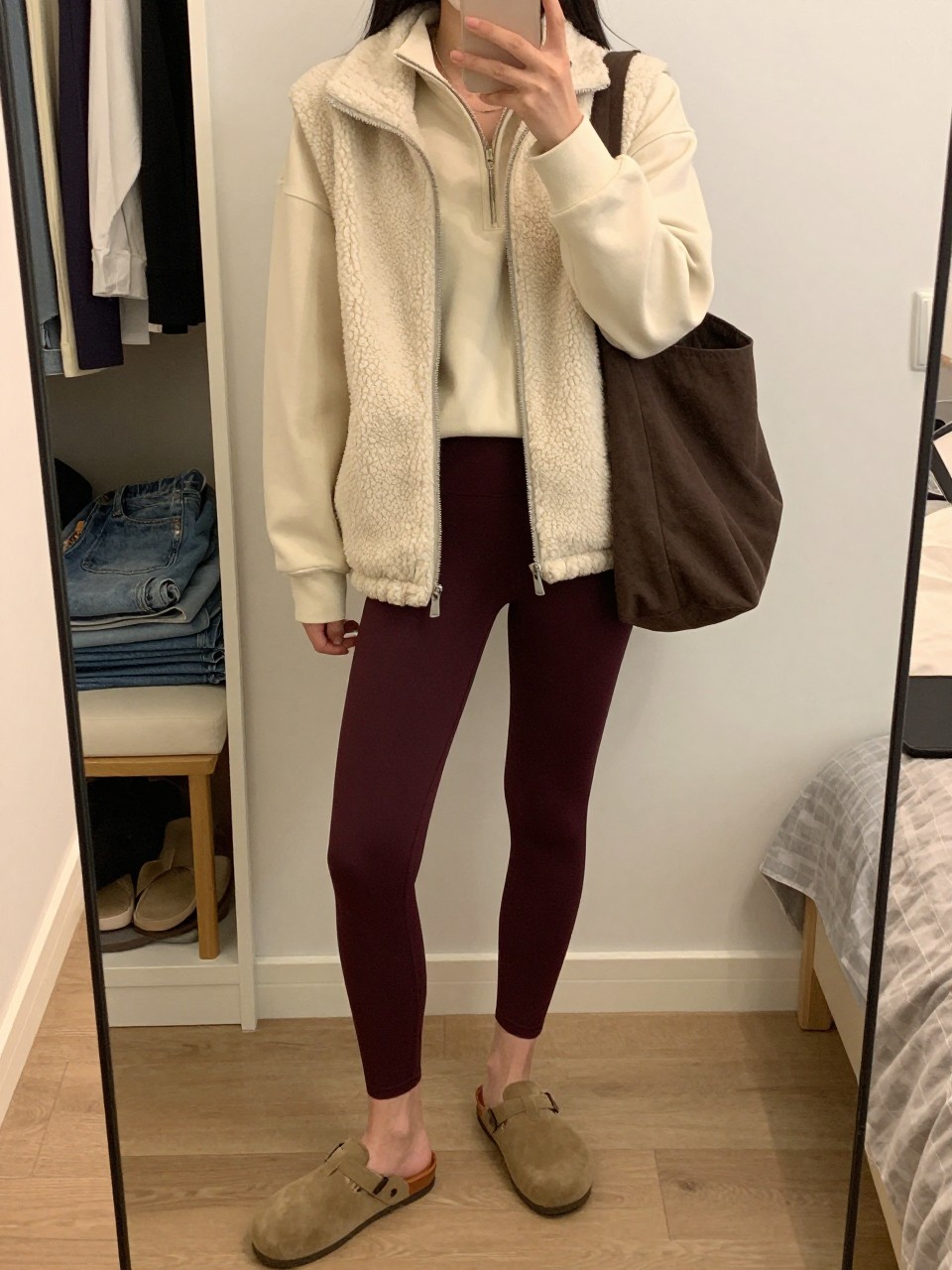 Mirror selfie of a woman wearing deep burgundy leggings, a cream quarter-zip sweatshirt, an ivory fleece vest, tan suede slip-on clogs, and a dark brown canvas tote bag, clearly showing the exact clothing items, colors, fabrics, and accessories listed, holding a phone in front of a feminine-themed bedroom mirror, cropped at the neck so the face does not show, relaxed casual selfie pose, warm moody lighting, open closet with clothes visible and folded denim jeans on a bench, casual uncluttered environment, latest iPhone photo quality.