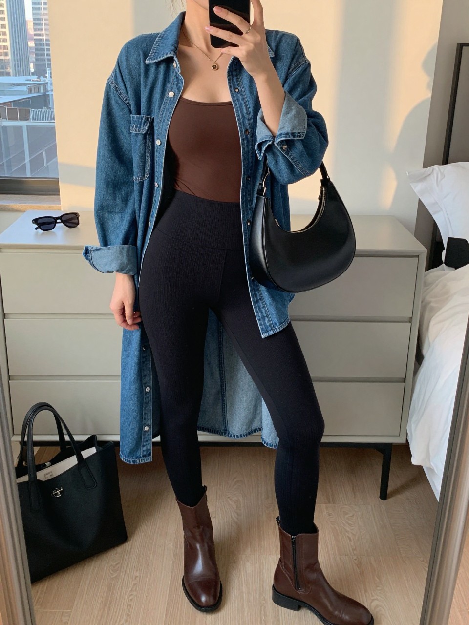Mirror selfie of a woman wearing black ribbed leggings, a fitted chocolate-brown tank top, a longline medium-wash denim shirt worn open, brown leather ankle boots, and a black crescent shoulder bag, clearly showing the exact clothing items, colors, fabrics, and accessories listed, holding a phone in front of a stylish city apartment mirror, cropped at the neck so the face does not show, relaxed casual selfie pose, evening golden-hour lighting, sunglasses placed on a dresser and a tote bag resting on the floor, natural lived-in composition, latest iPhone photo quality.