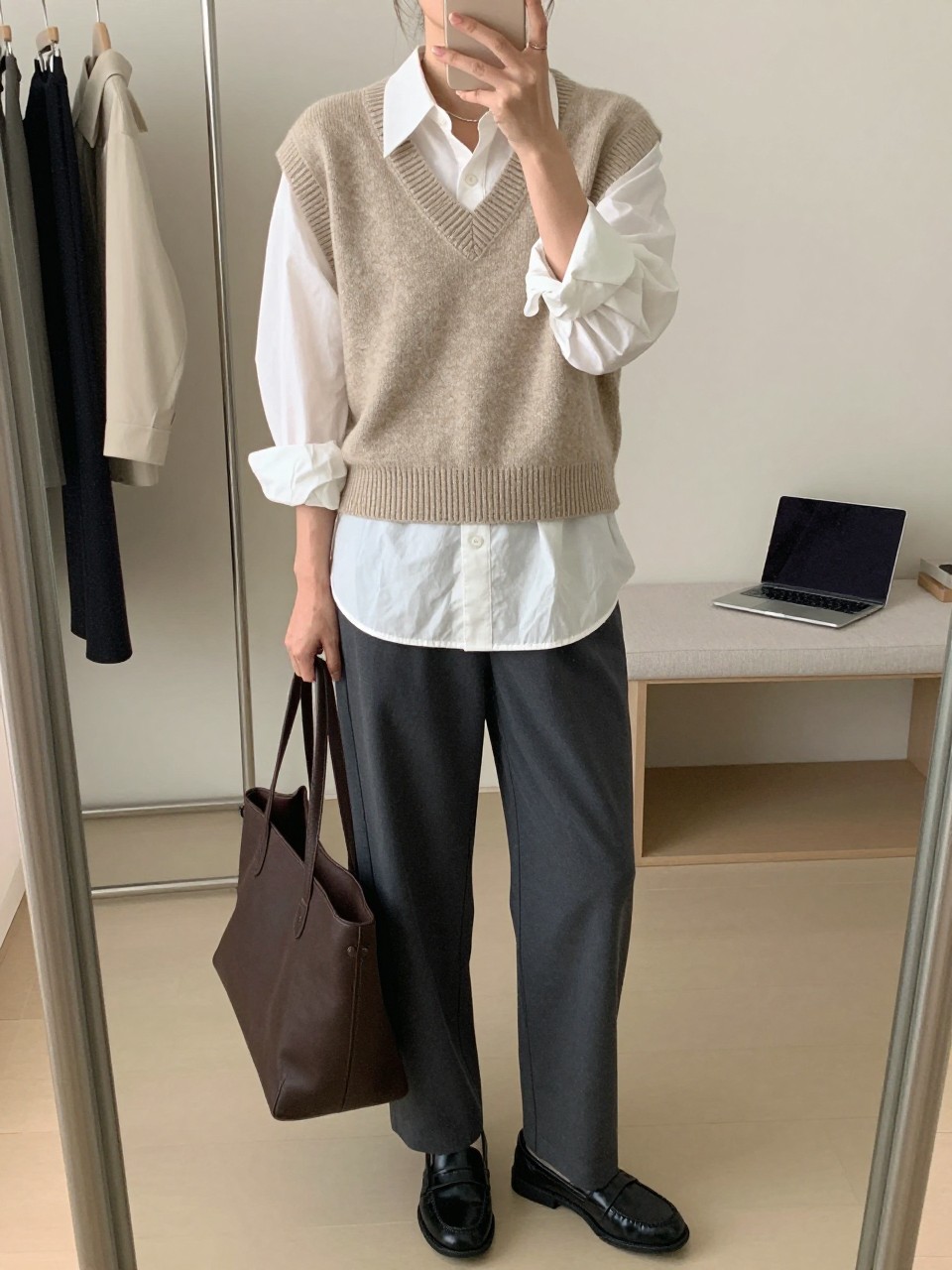 Mirror selfie of a woman wearing an oatmeal V-neck sweater vest layered over an oversized white poplin button-front shirt, charcoal-grey straight-leg trousers, black leather loafers, and a structured dark brown tote bag, clearly showing the exact clothing items, colors, fabrics, and accessories listed, holding a phone in front of a minimal dressing room mirror, cropped at the neck so the face does not show, relaxed casual selfie pose, natural window light, a clothing rack with garments behind and a laptop on a bench, polished yet natural, latest iPhone photo quality.