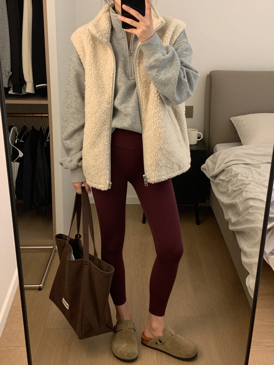 Mirror selfie of a woman wearing a cream fleece vest over an oversized heather-grey quarter-zip sweatshirt, deep burgundy high-waisted leggings, tan suede clogs, and a dark brown canvas tote bag, clearly showing the exact clothing items, colors, fabrics, and accessories listed, holding a phone in front of a cozy bedroom mirror setup, cropped at the neck so the face does not show, relaxed casual selfie pose, warm moody lighting, an open closet with clothes visible and a coffee mug on a bedside table, casual and realistic, latest iPhone photo quality.