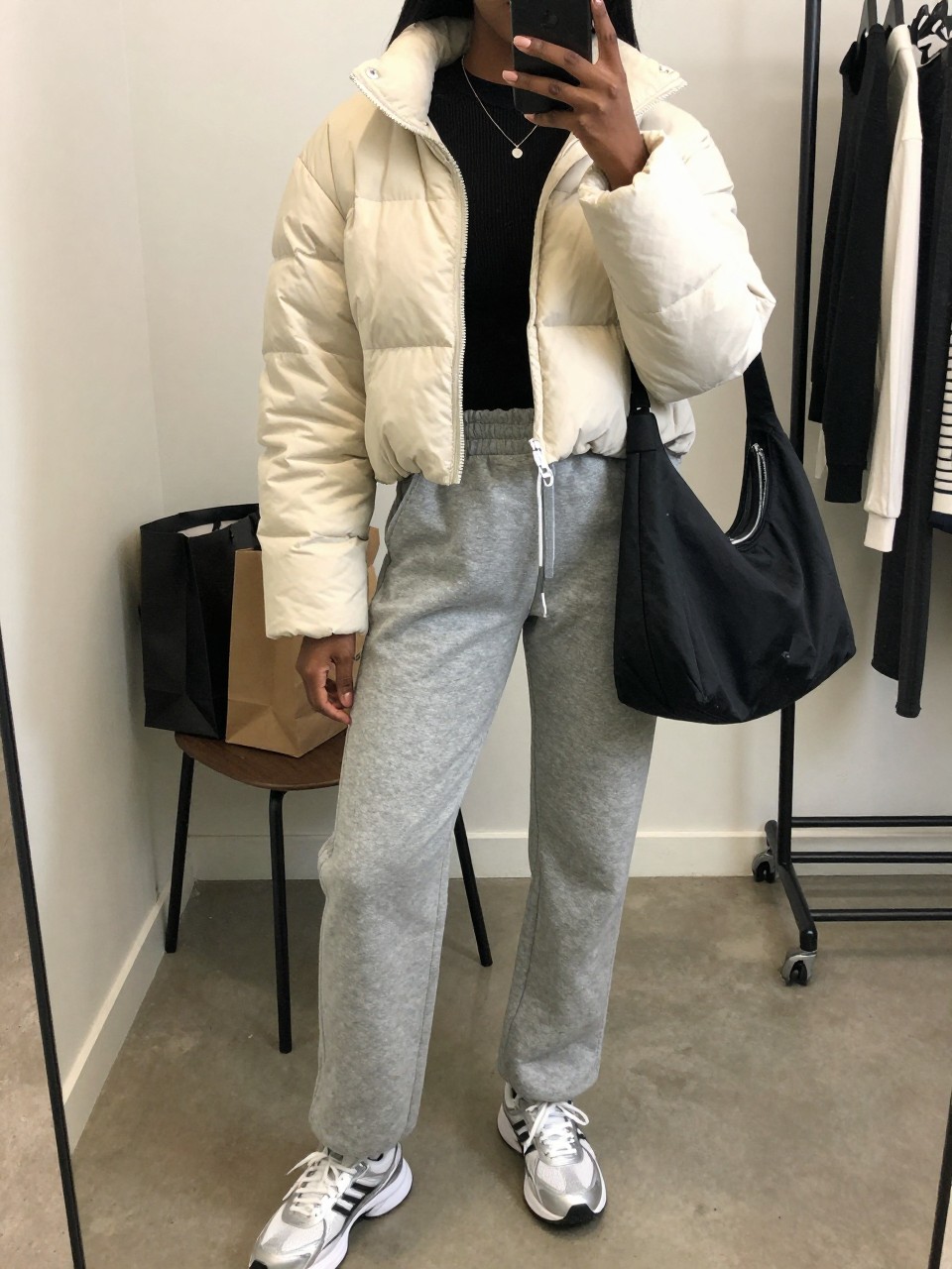 Mirror selfie of a Black woman wearing an ivory cropped quilted puffer jacket, fitted black ribbed long-sleeve top, heather-grey straight-leg sweatpants, white and silver retro sneakers, and a black nylon shoulder bag, clearly showing the exact clothing items, colors, fabrics, and accessories listed, holding a phone in front of a fashion studio mirror corner, cropped at the neck so the face does not show, relaxed casual selfie pose, soft indoor lighting, shopping bags beside a chair and a clothing rack with garments behind, believable casual setting, latest iPhone photo quality.