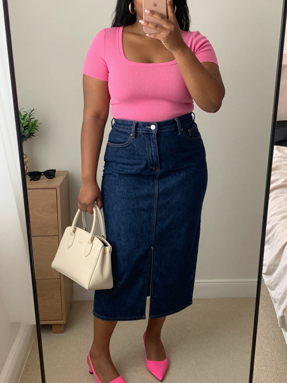 Mirror selfie of a black plus size woman wearing a dark indigo high-waisted denim midi skirt with a front walking slit, a fitted hot pink short-sleeve square-neck knit top, hot pink pointed slingback flats, a structured cream handbag, and chunky gold hoop earrings, clearly showing the exact clothing items, dark denim, hot pink knit, front slit, footwear, and accessories listed, holding a phone in front of a mirror, cropped at the neck so her face is hidden, relaxed playful selfie stance, in a cozy bedroom mirror setup with soft indoor lighting, sunglasses placed on a dresser and a small plant nearby, realistic casual dinner-date outfit, latest iPhone photo quality.