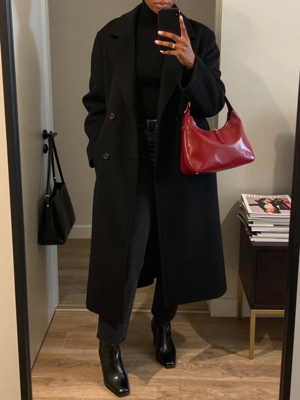 Mirror selfie of a Black woman wearing a long black wool coat, fitted black ribbed turtleneck, black straight-leg jeans, black leather square-toe ankle boots, and a vivid cherry-red shoulder bag, clearly showing the exact clothing items, colors, fabrics, and accessories listed, holding a phone in front of a stylish city apartment mirror, cropped at the neck so the face does not show, relaxed confident selfie pose, warm moody lighting, a handbag hanging on a door handle and a stack of fashion magazines on a side table, sleek and natural, latest iPhone photo quality.